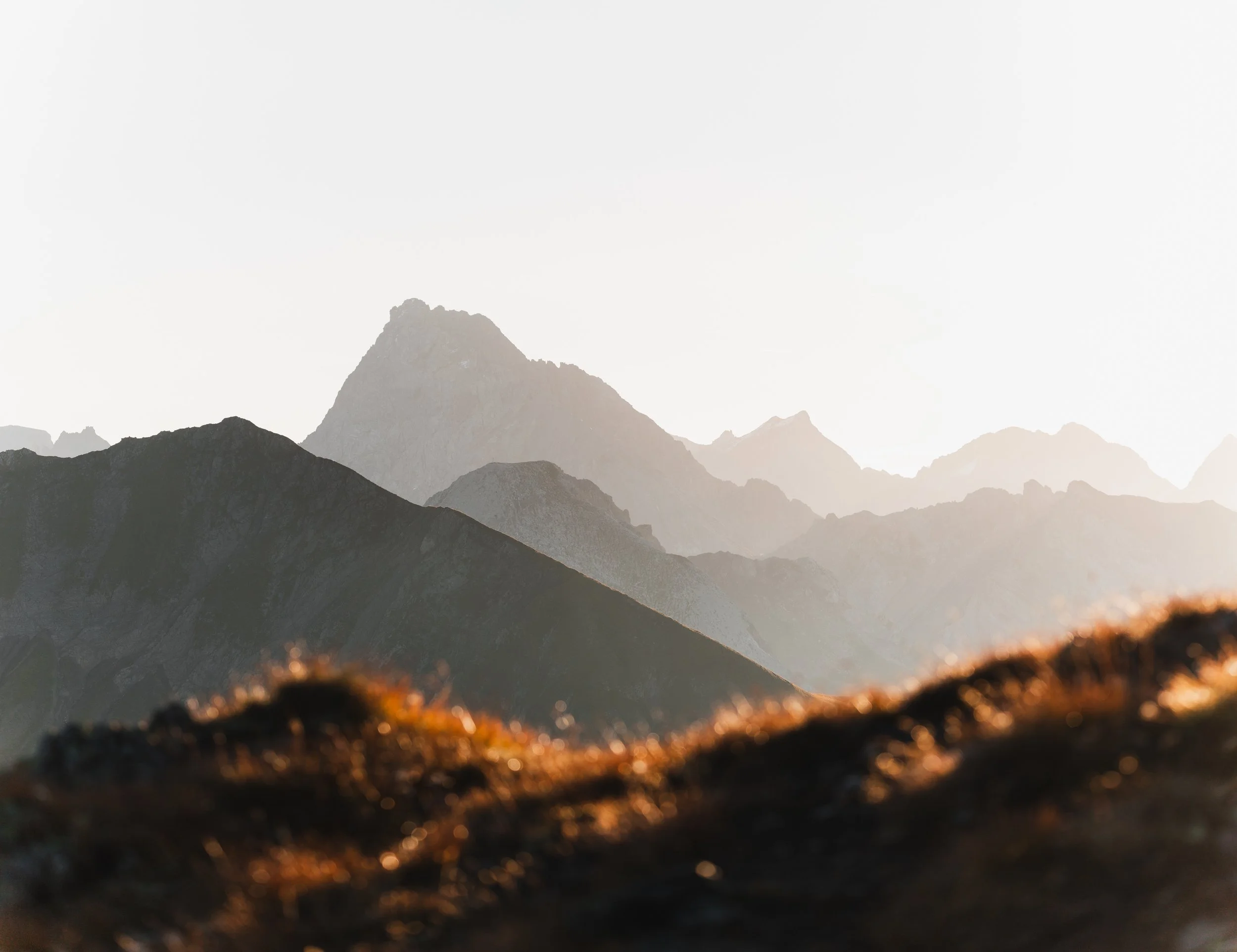Berglandschaft mit mehreren Bergen bei Sonnenaufgang oder Sonnenuntergang, im Vordergrund trockenes Gras.