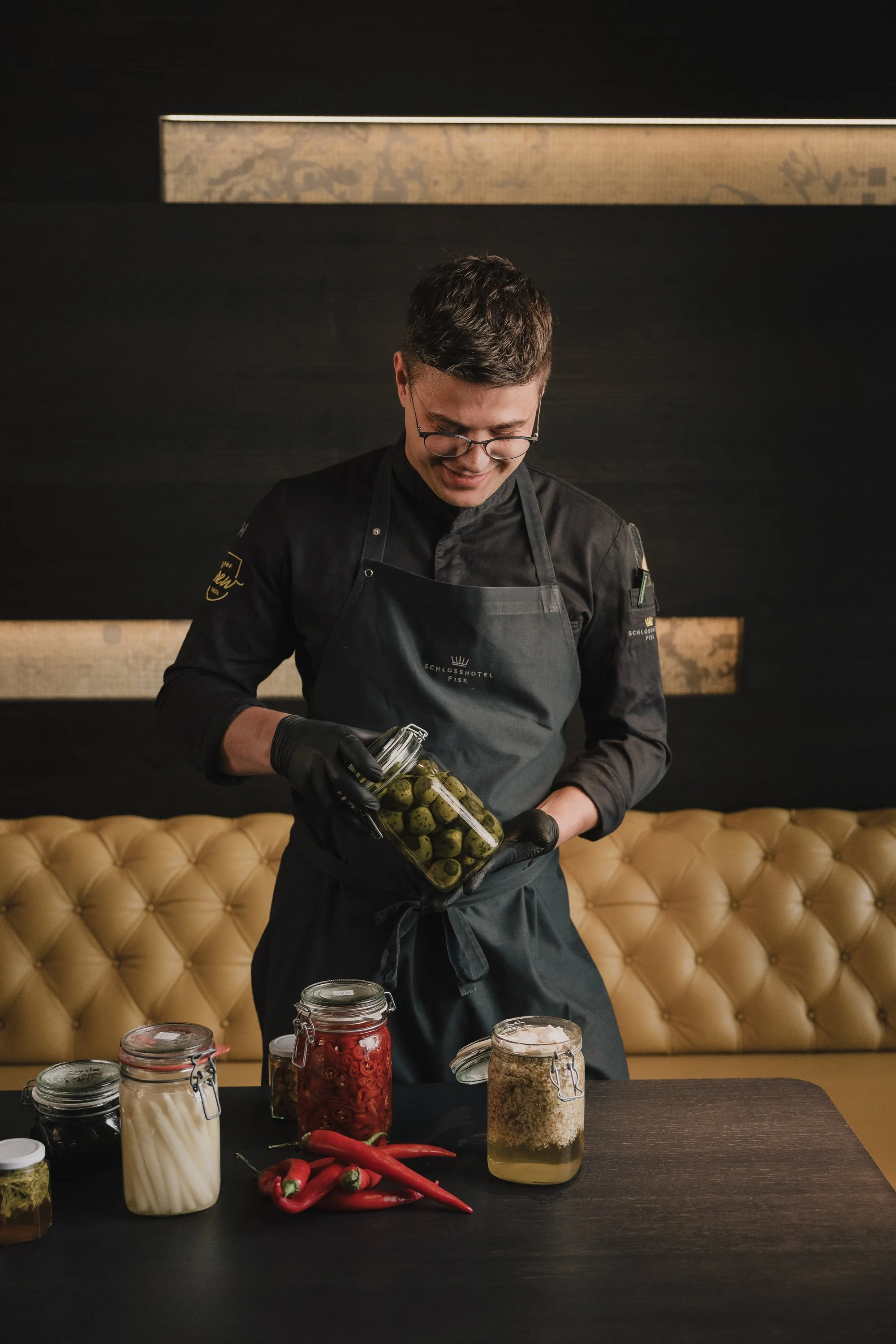 Ein junger Koch in schwarzer Kochuniform und Schürze, der in einer modern eingerichteten Küche mit dunklem Hintergrund und bequemer Ledersitzbank im Hintergrund eingelegte grüne Oliven in einem Glas hält.
