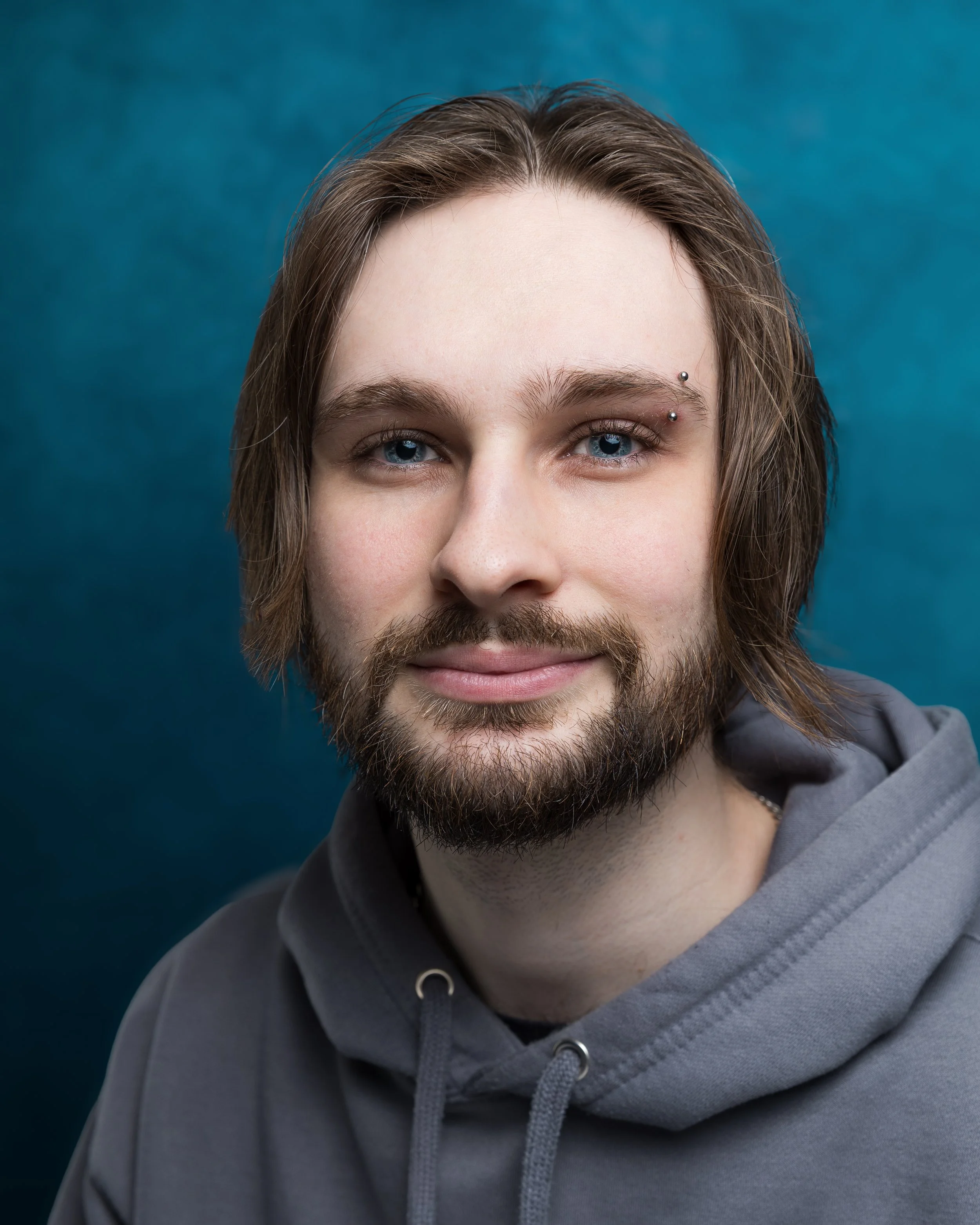 A young man with long brown hair, glasses, and a beard, wearing a white t-shirt with blue designs, posing against a gray background.