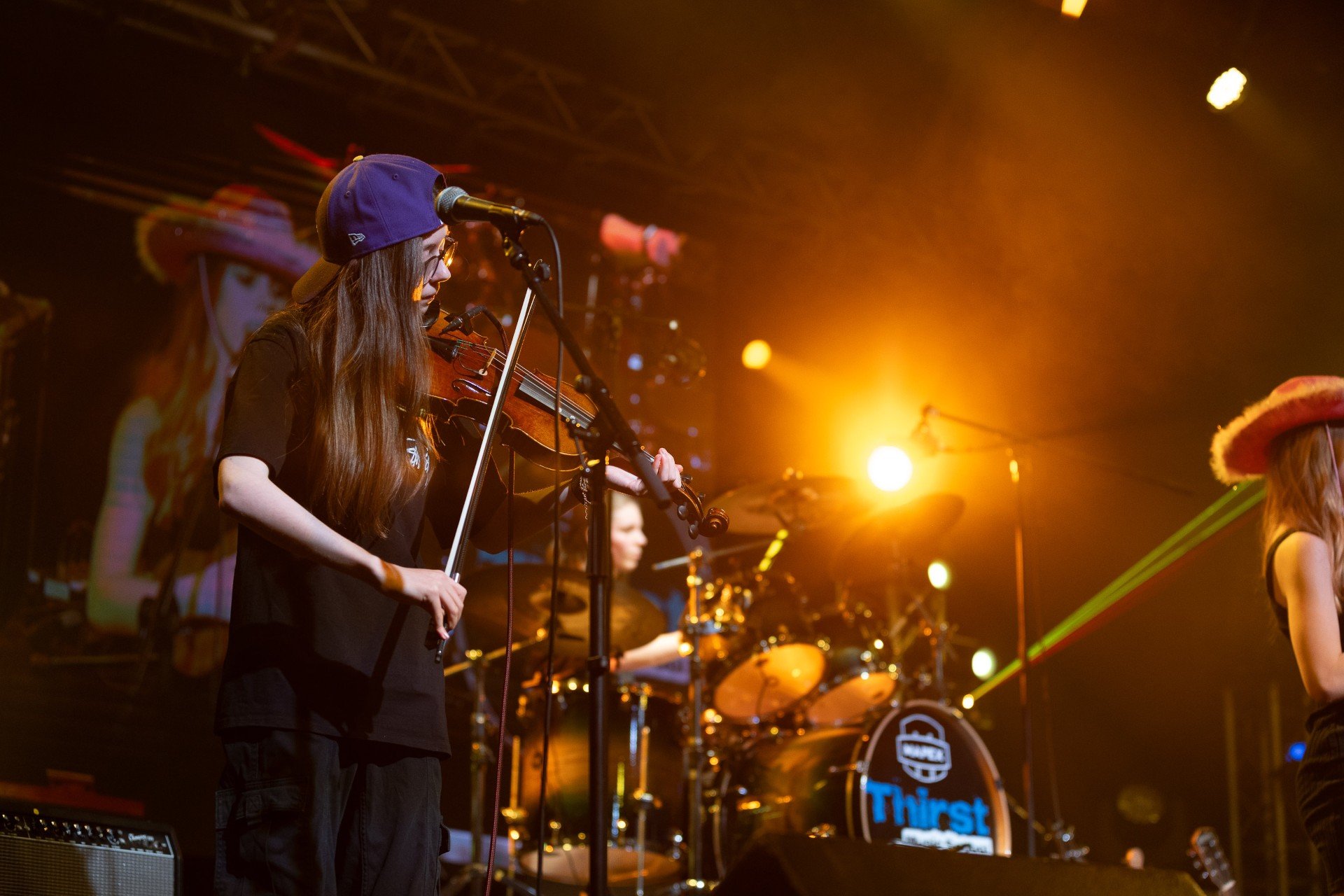 A female musician with long hair, glasses, a blue cap, playing a violin on stage with yellow and orange stage lights, and a drummer in the background.