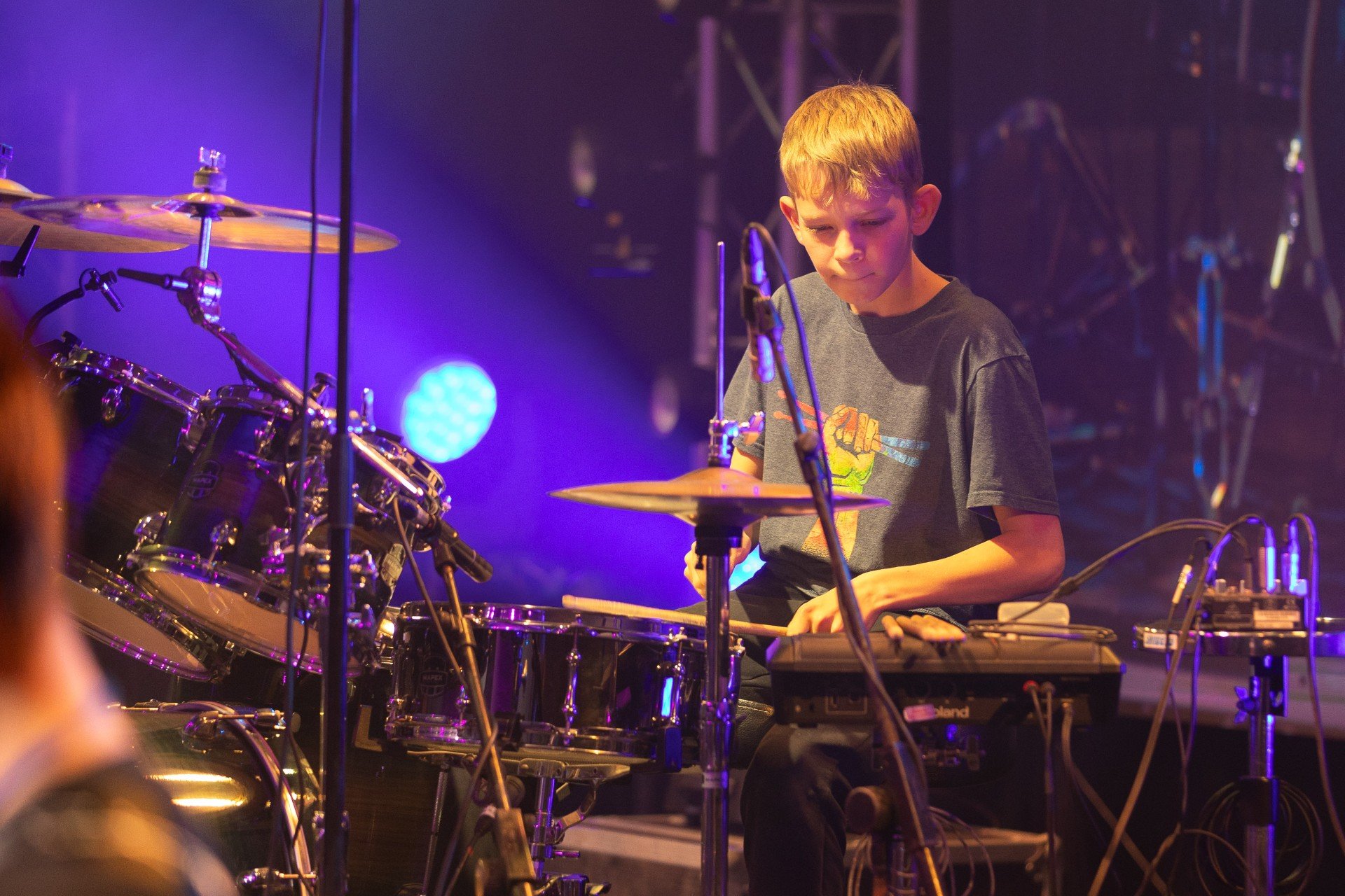 Young boy playing drums and electronic keyboard on stage during a performance, illuminated by blue and purple stage lights.