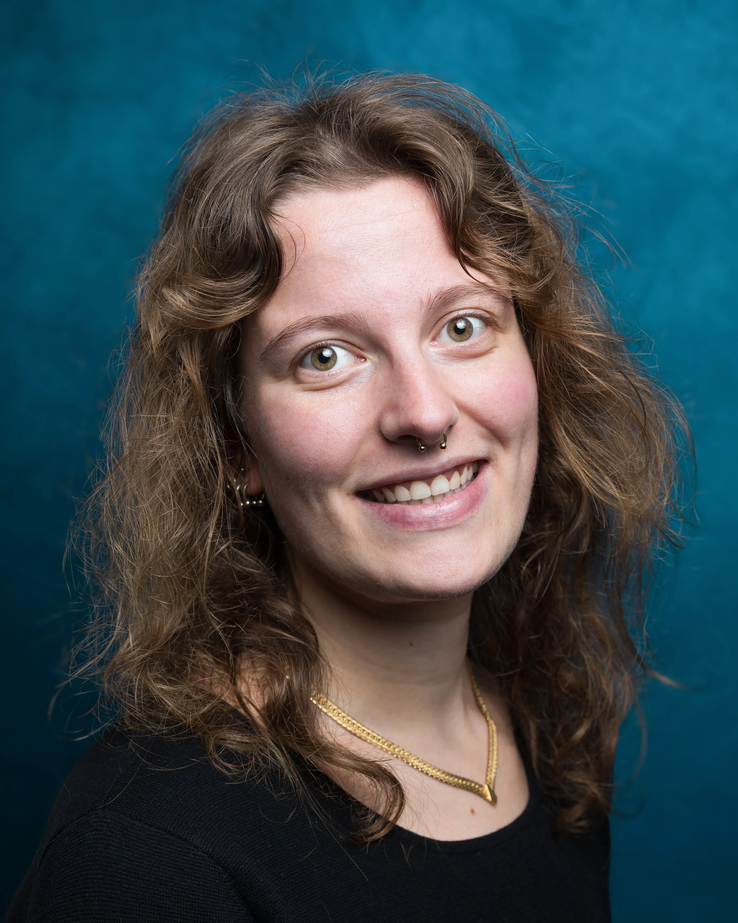 A woman with light skin, long wavy brown hair, and blue eyes, smiling slightly, wearing a septum piercing, a patterned top, a choker necklace, and a silver chain. She is posing against a dark textured background.