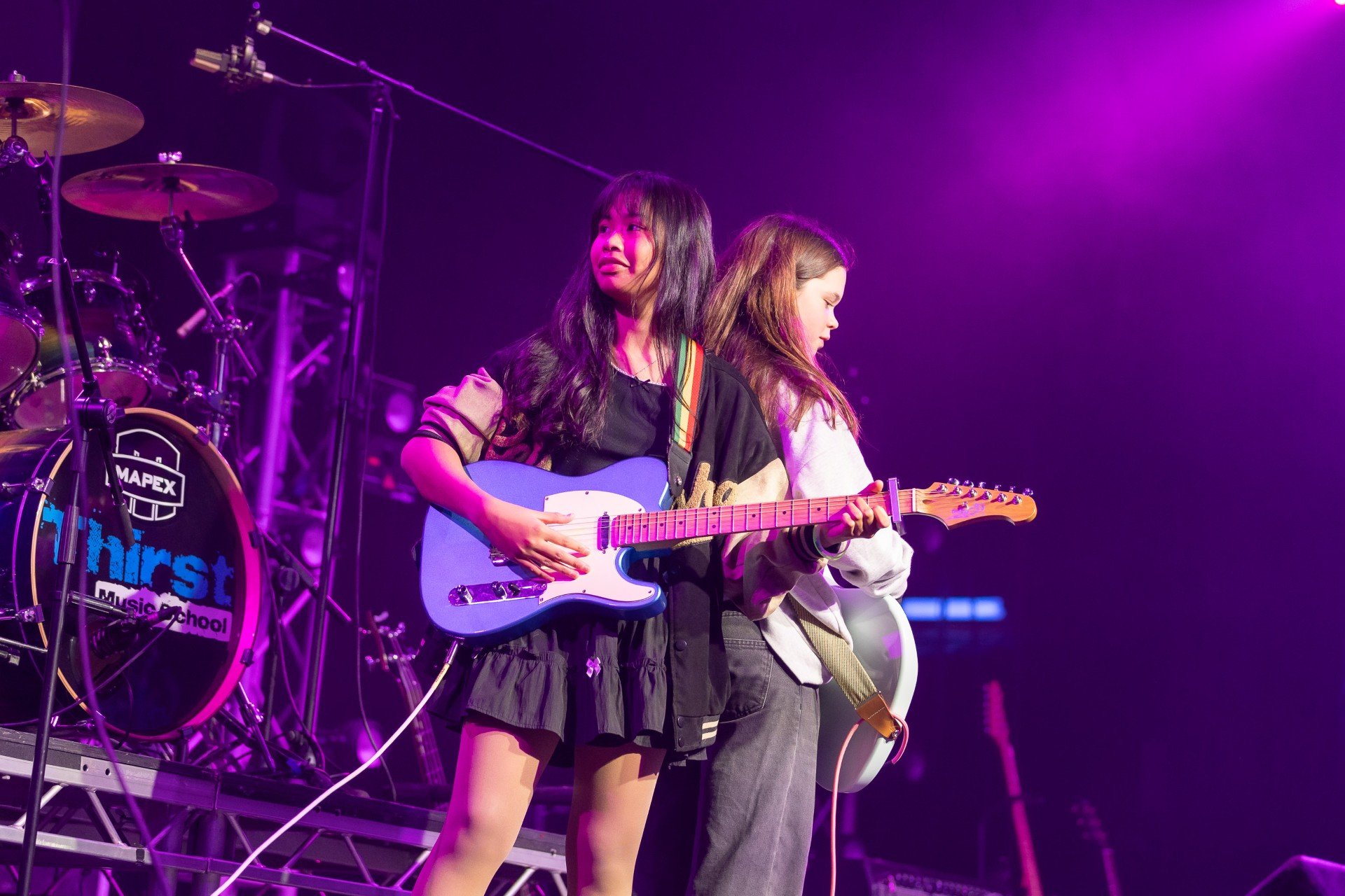 Two young women playing guitars on stage under purple and pink lighting, with drums and stage equipment in the background.