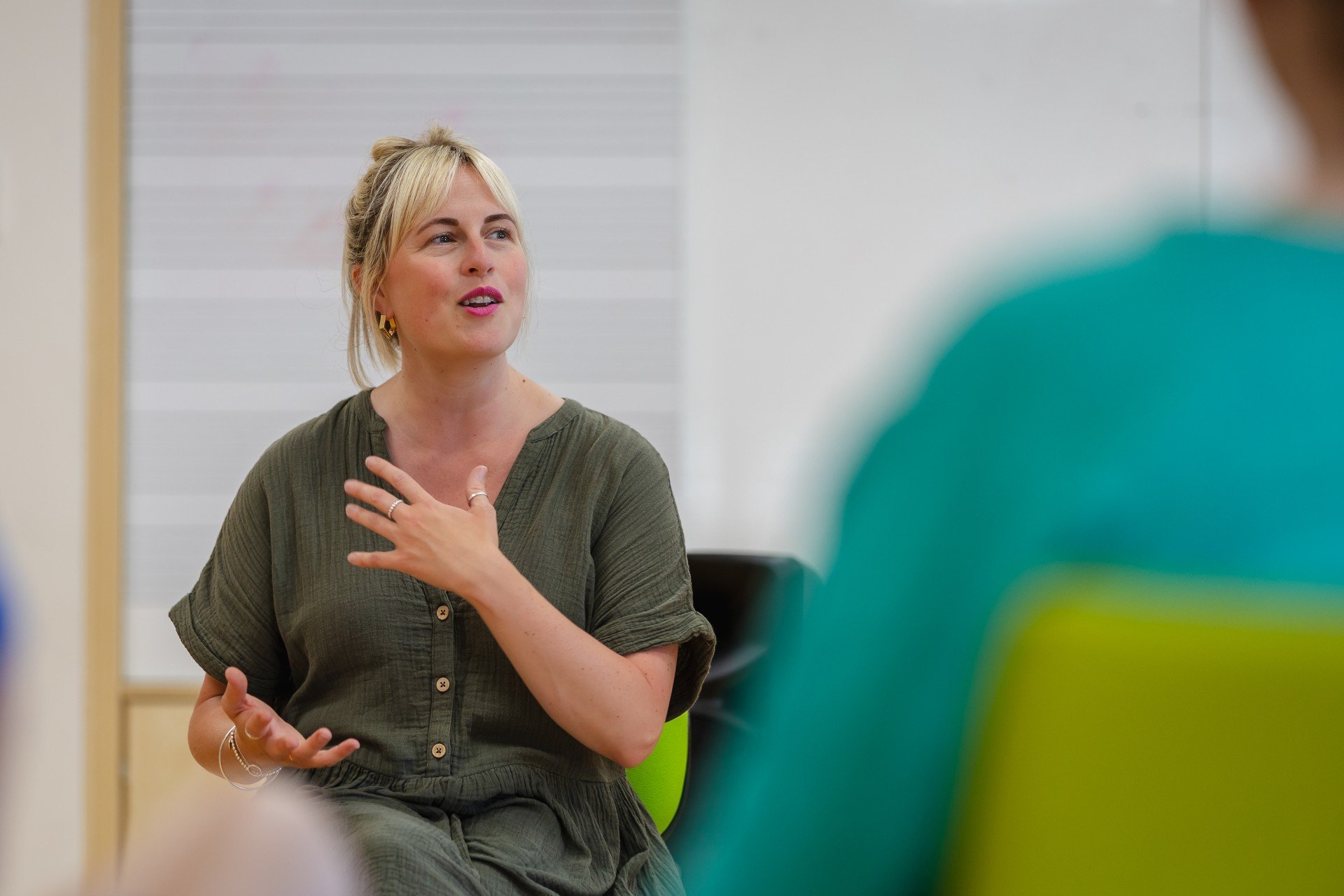 A woman with blonde hair and earrings speaking in a classroom or meeting room, with a whiteboard in the background.