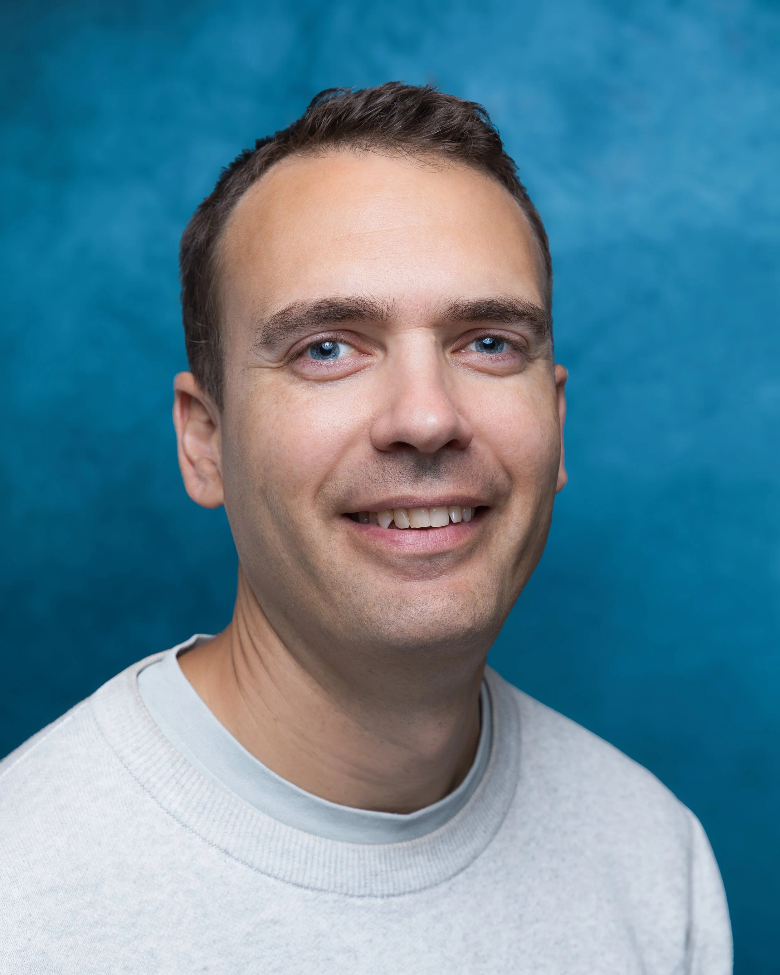 Portrait of a smiling man with dark hair and blue eyes, wearing a black jacket over a blue shirt, against a dark, textured background.