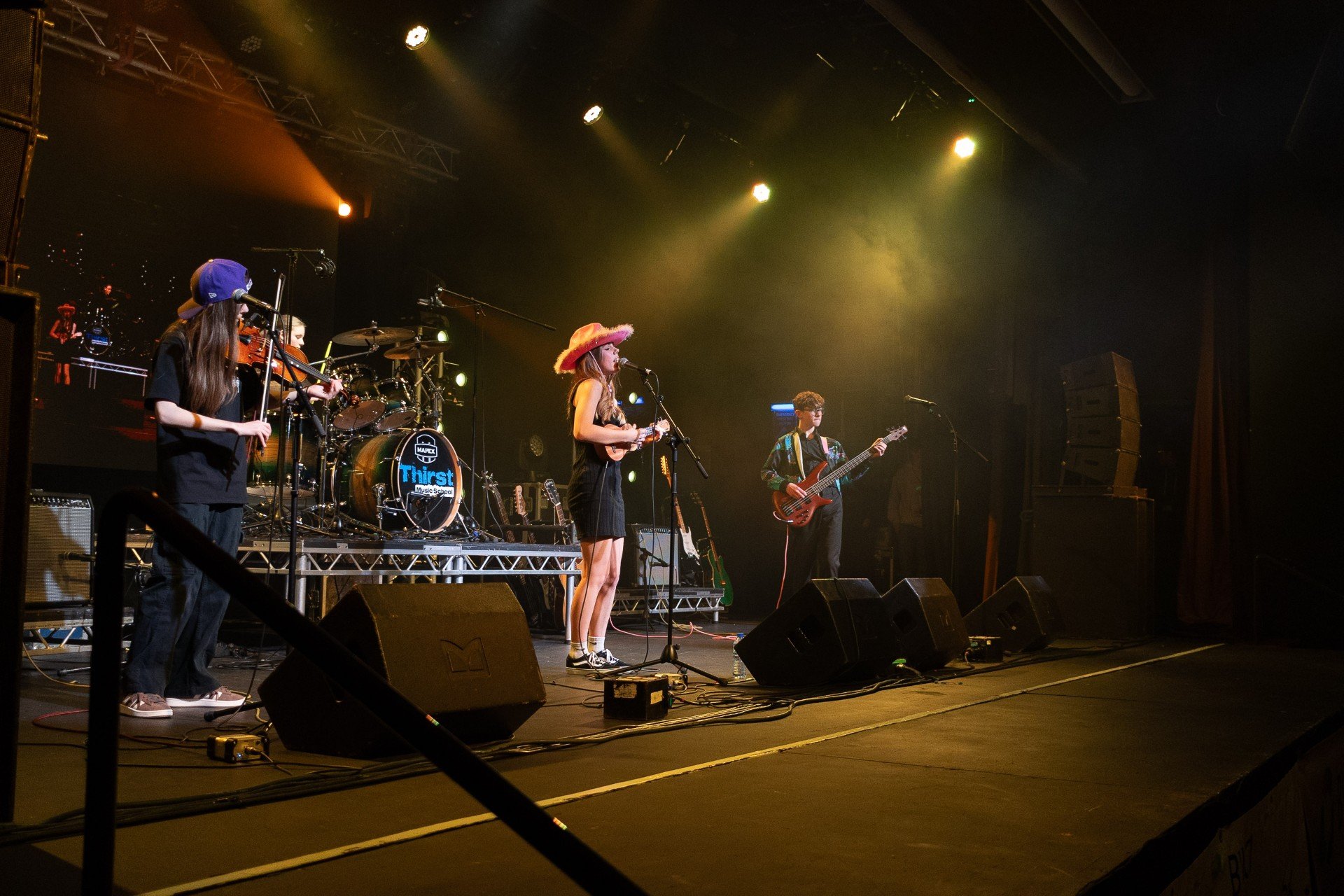 A live musical performance on stage featuring four musicians, including a violinist, a singer with a ukulele, a bassist, and a drummer, with colorful stage lighting and a large screen in the background.