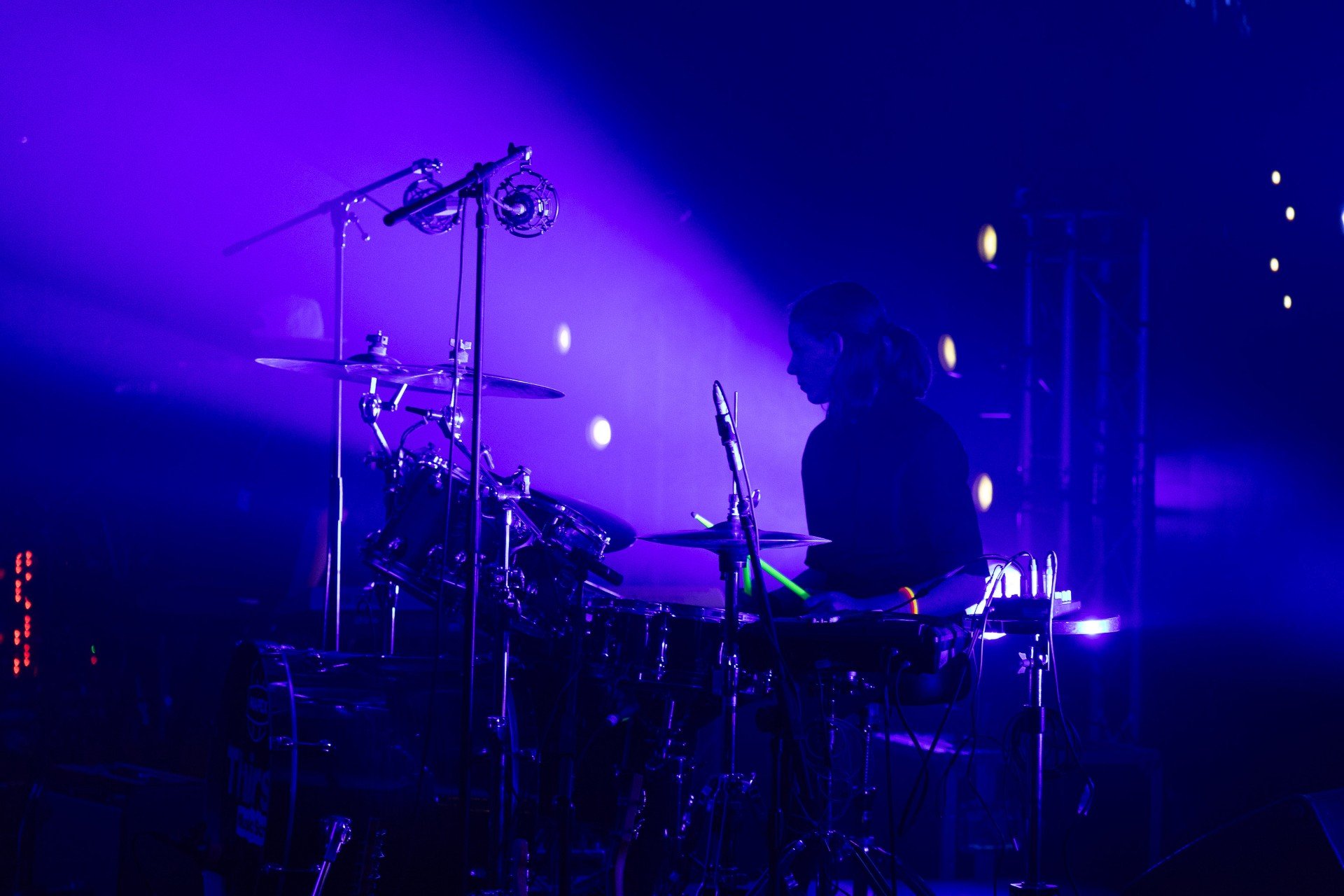 A person playing drums on stage during a concert, illuminated by purple and blue stage lights.