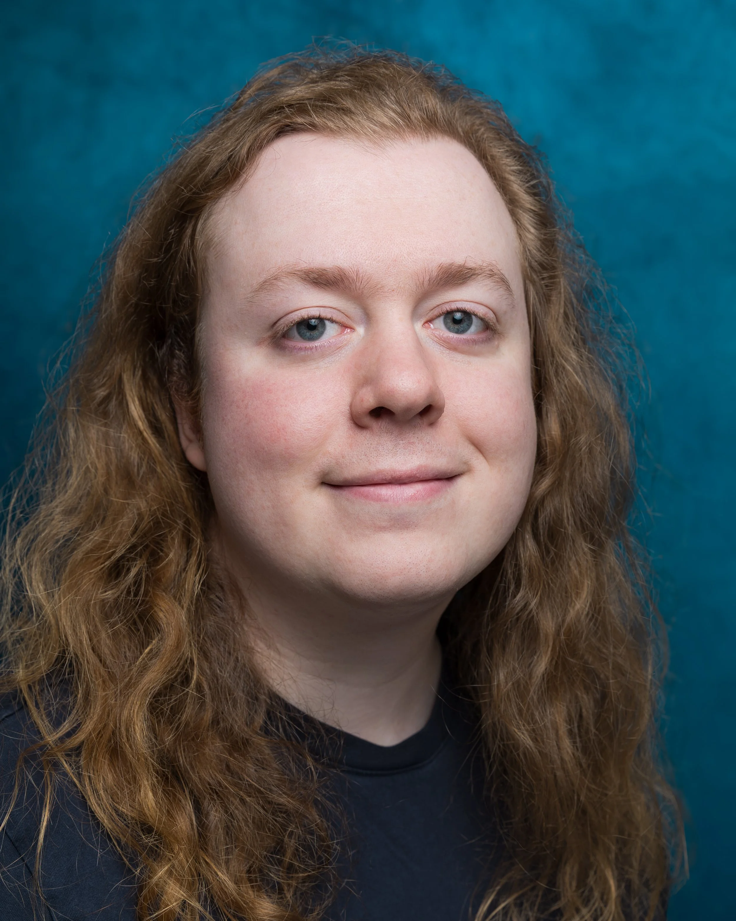 A young man with long, wavy red hair, fair skin, and blue eyes, smiling and wearing a black shirt against a dark, textured background.