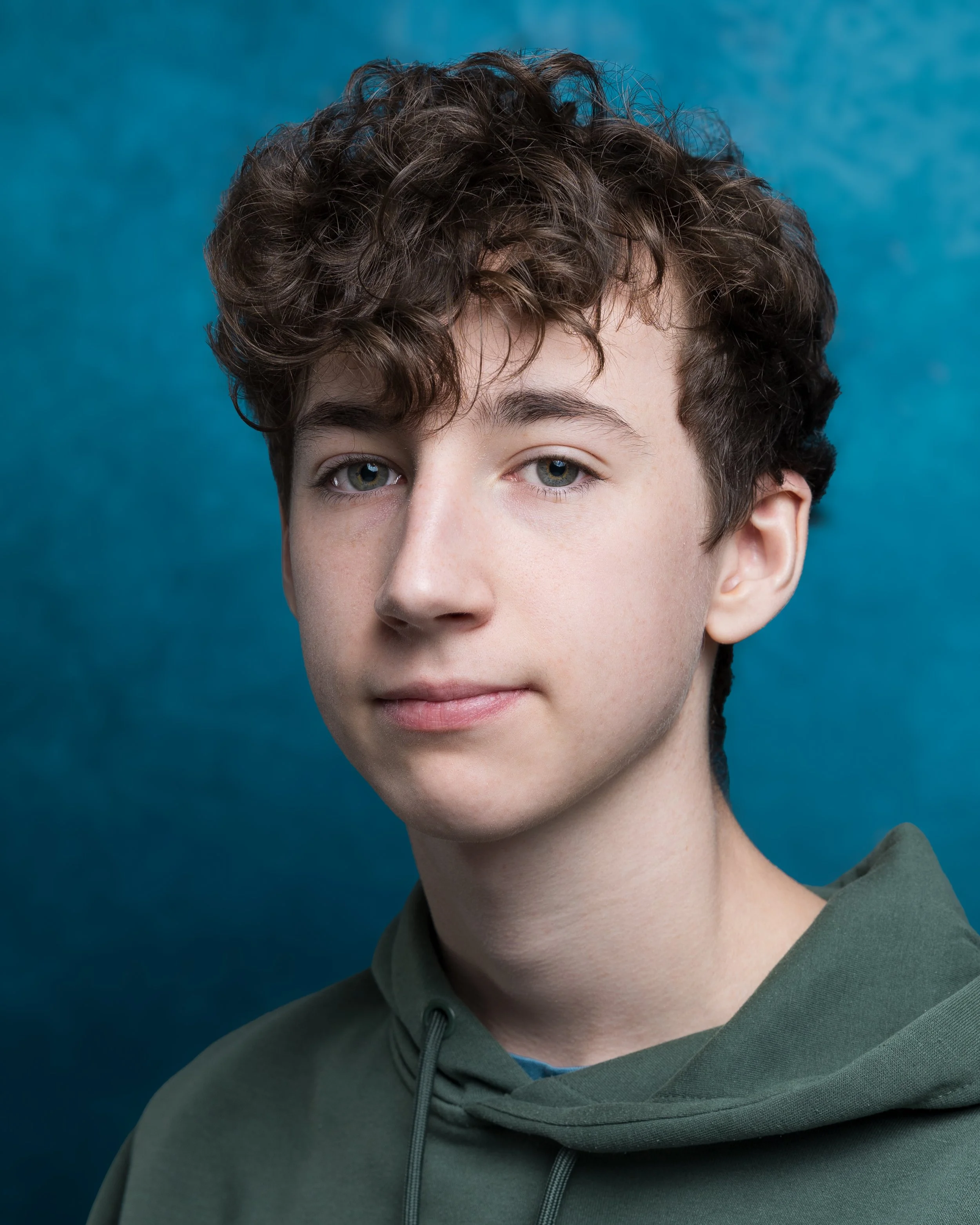 A teenage boy with curly brown hair and light skin, wearing a blue hoodie with white text, posing in front of a dark purple background.