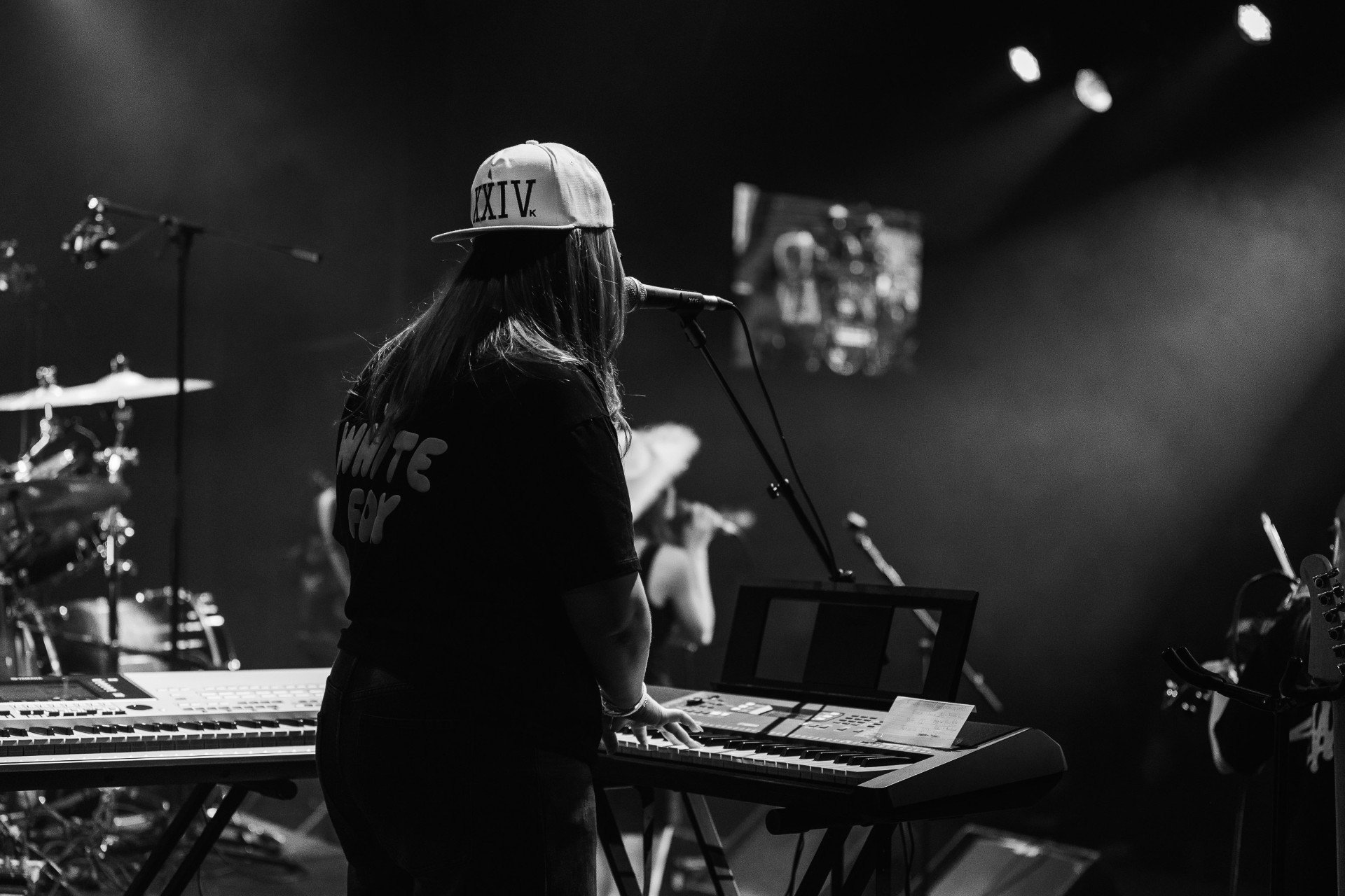 A female musician wearing a baseball cap and a T-shirt, playing a keyboard on stage during a live music performance. There are drums and other equipment visible in the background.