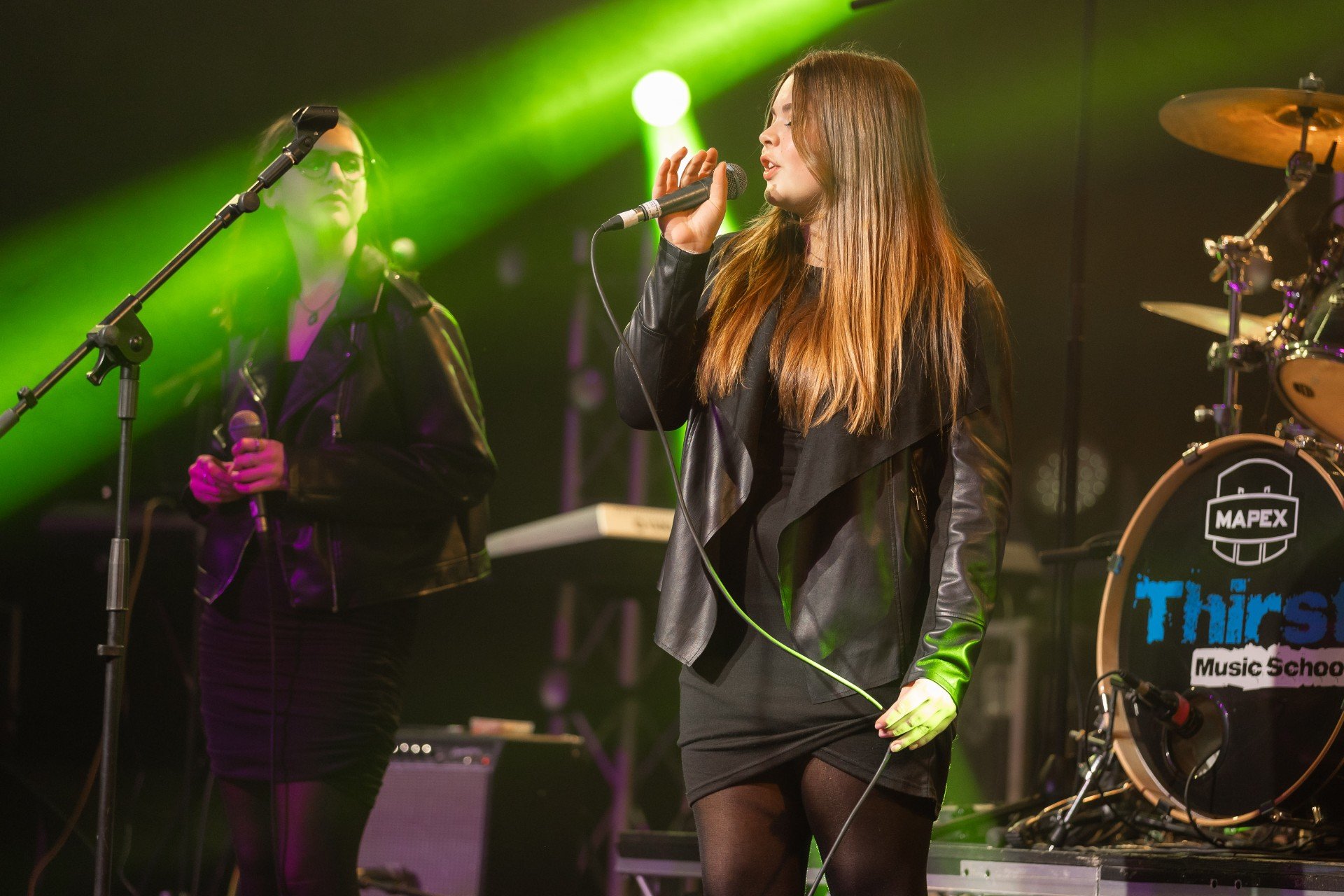 Two women performing on stage with musical instruments, one singing into a microphone and the other holding a microphone, with green stage lights and a drum set in the background.