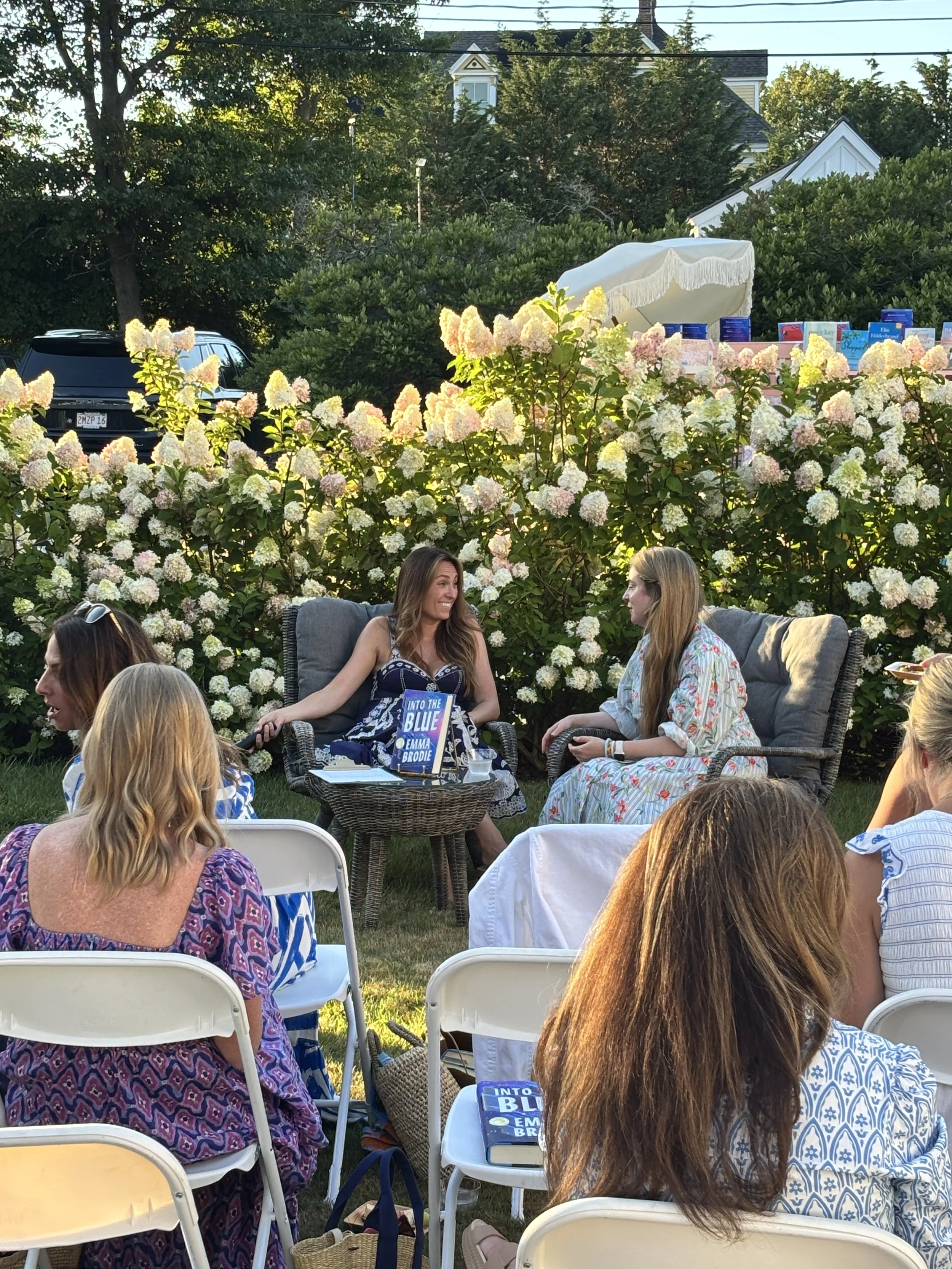 People gathered outdoors at a social event, sitting on chairs and couches, with a backdrop of lush green bushes and trees, and a house with a pitched roof and windows in the background.