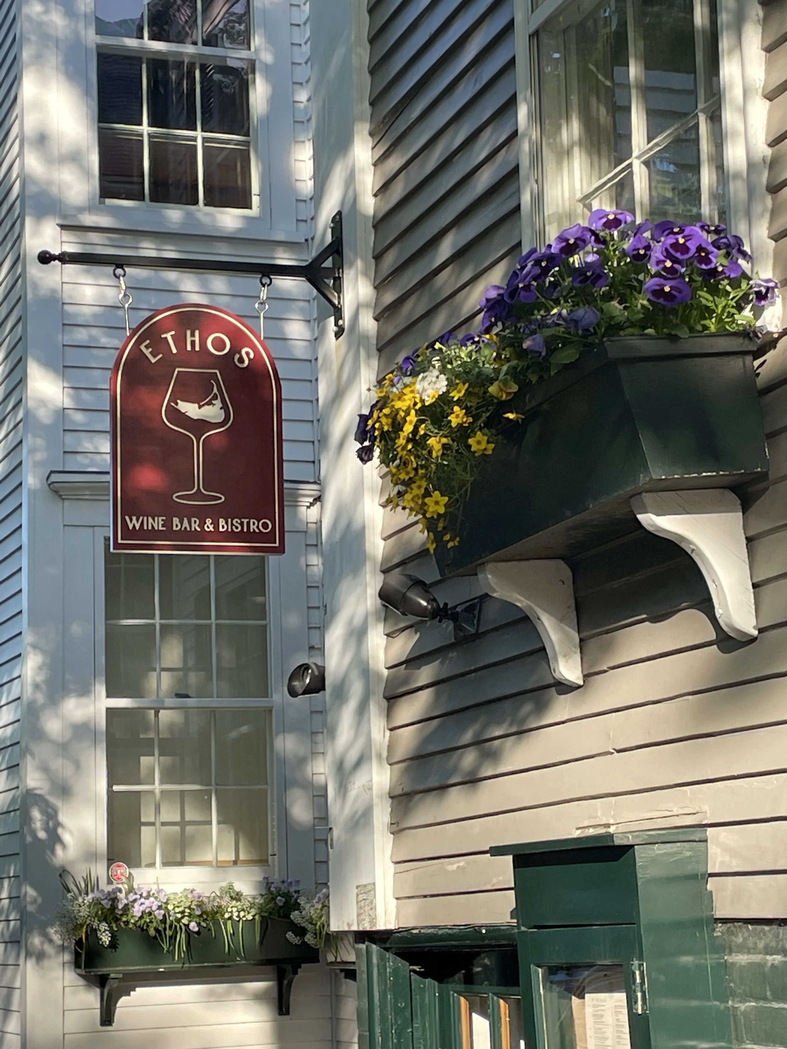 Red sign for a wine bar and bistro named Ethos, hanging on a gray building with flower boxes beneath windows; purple and yellow flowers in the boxes.