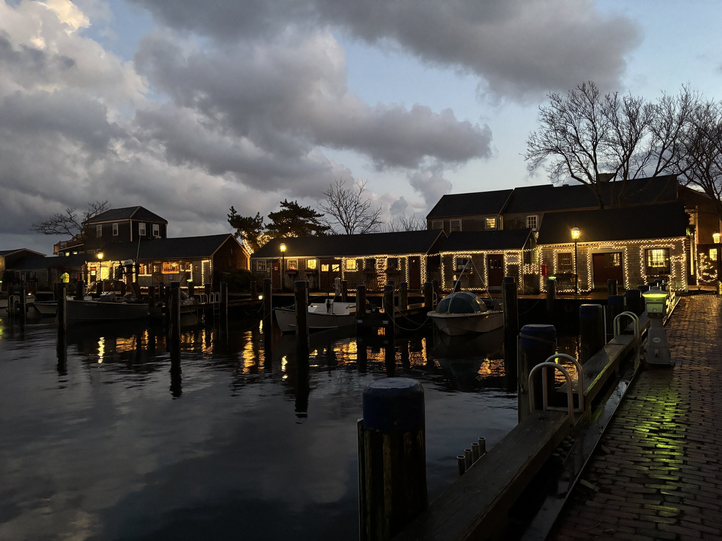 Nantucket harbort scene with boats docked near buildings decorated with string lights at dusk, reflecting on water, cloudy sky overhead.