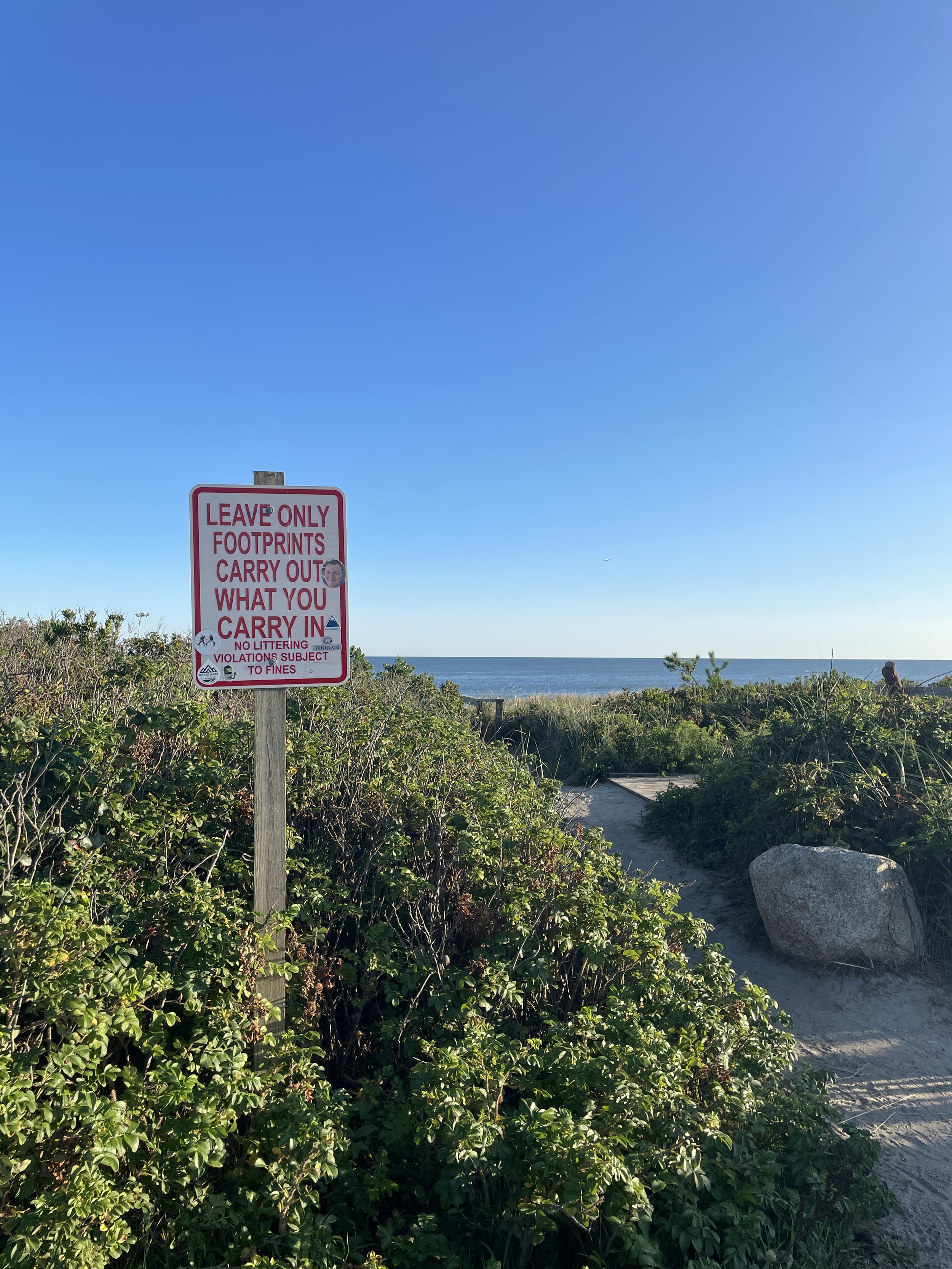 A sign surrounded by greenery with the ocean in the background. The sign reads: 'Leave only footprints, carry out what you carry in, no littering, violations subject to fines.' The sky is clear and blue.