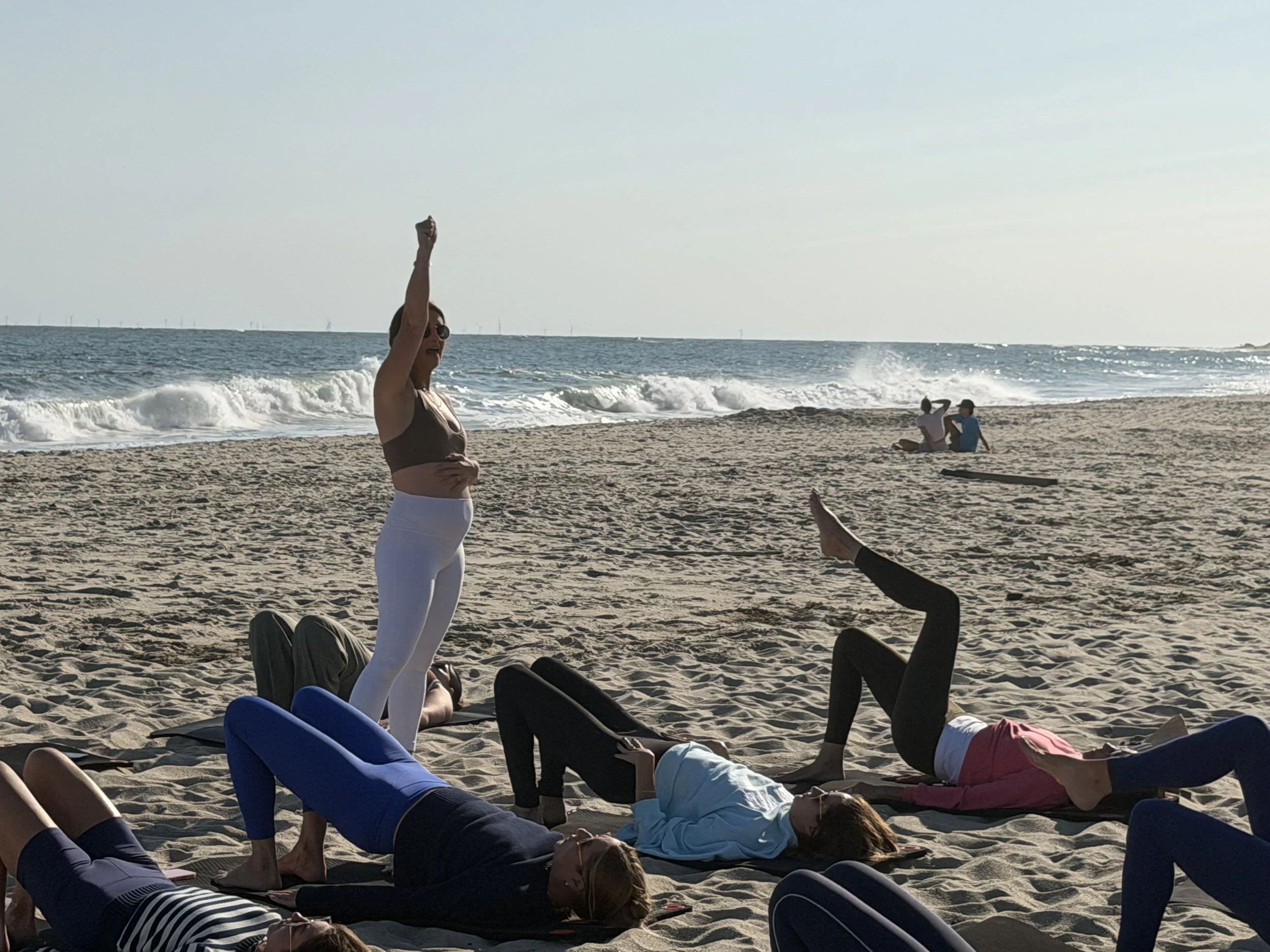 People practicing yoga on the beach with the ocean and wind turbines in the background during sunset.