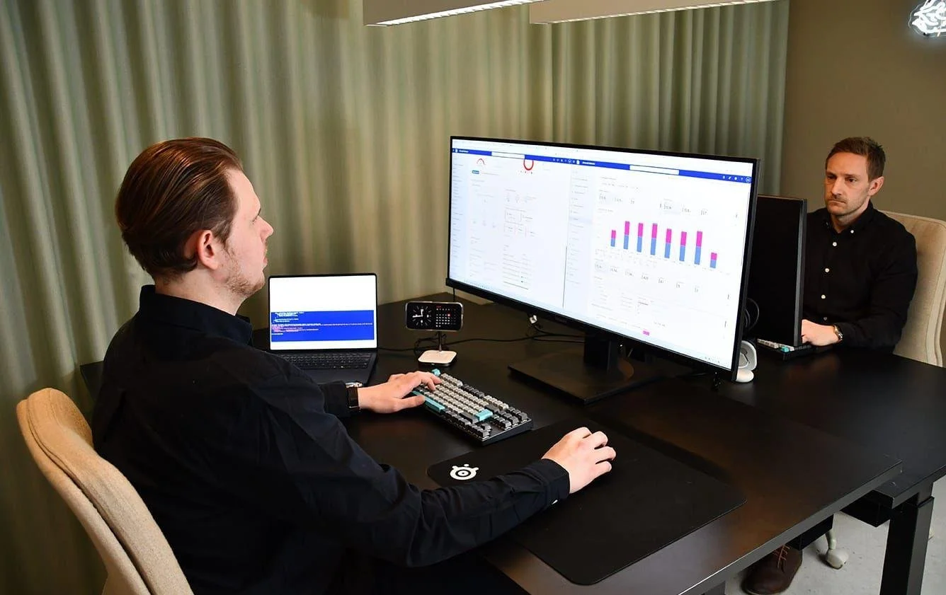 Två personer som arbetar vid ett mörkt bord, med datorskärmar som visar grafer och diagram, i ett mötesrum med gröna gardiner.