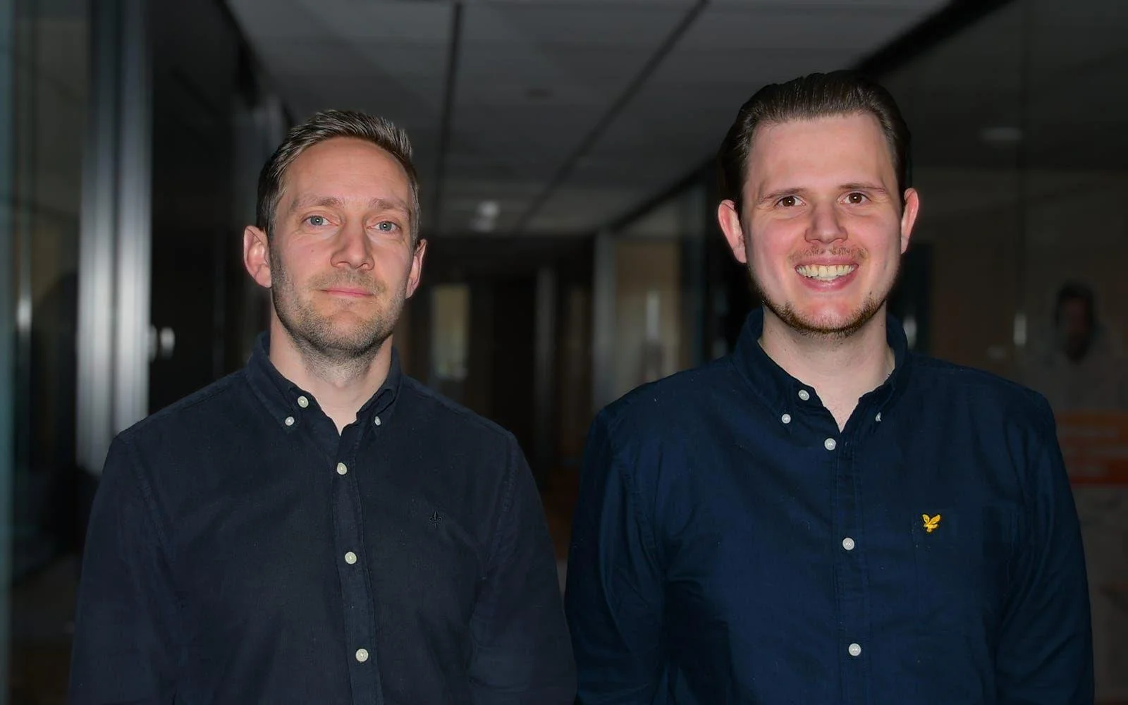 Två unga män står i en inomhusmiljö, båda bär mörk blå button-down skjorta. Den vänstra mannen har kort, ljus hår och ett allvarligt uttryck, den högra mannen har mörkare hår, ett brett leende och är yngre.