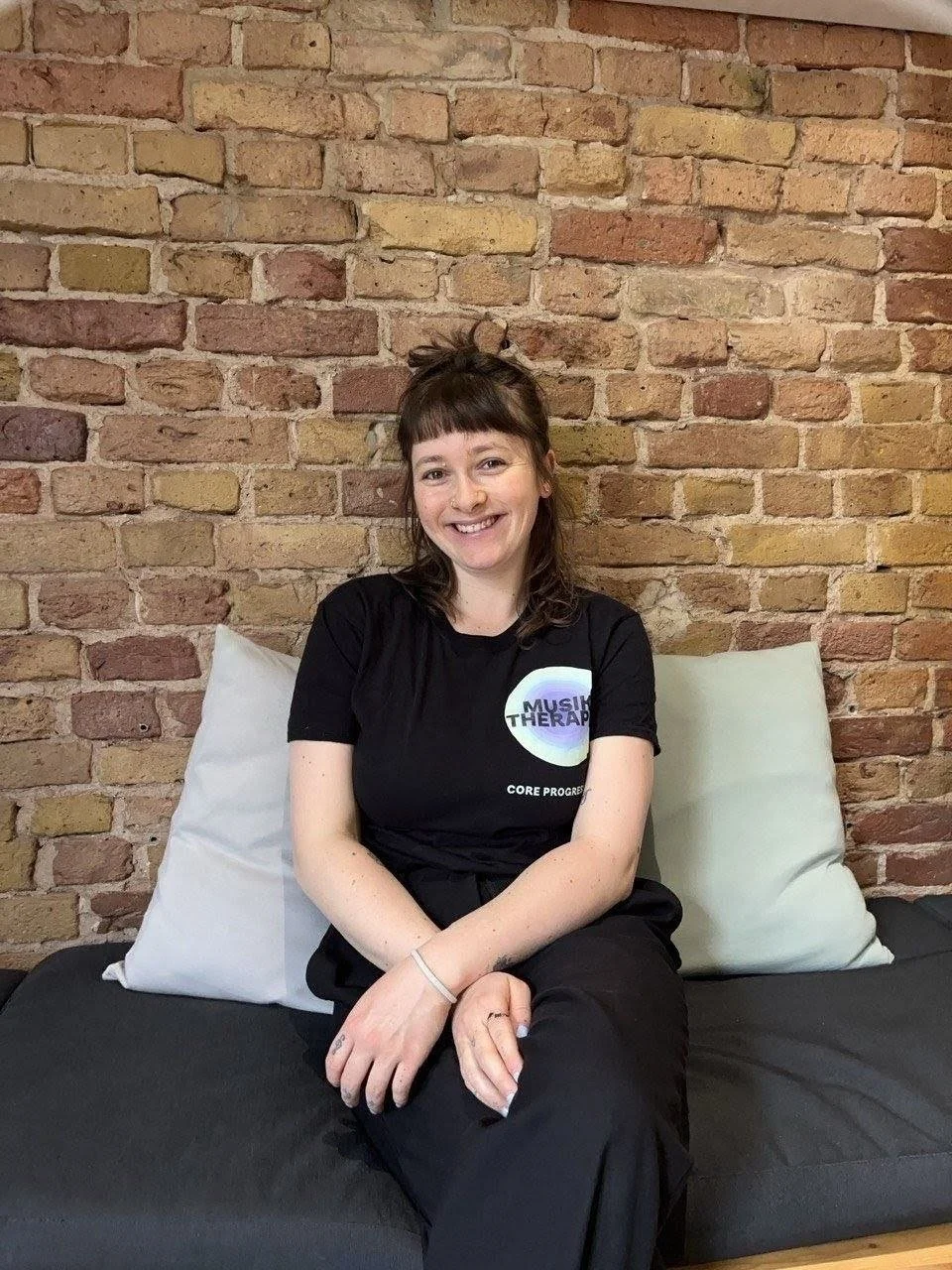 Junge Frau mit braunen Haaren und Schleife im Haar, lächelnd, sitzt auf einer Couch vor einer Ziegelwand, trägt ein schwarzes T-Shirt mit einem Musikkonzept-Logo.