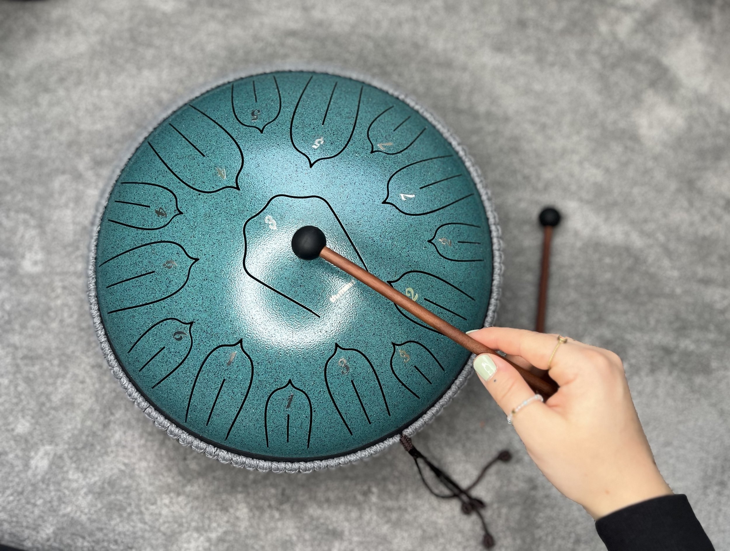 Nahaufnahme eines Handpan-Instruments auf einer schwarzen Oberfläche, mit einer Hand, die einen Klangstab hält, der auf das Instrument zeigt.