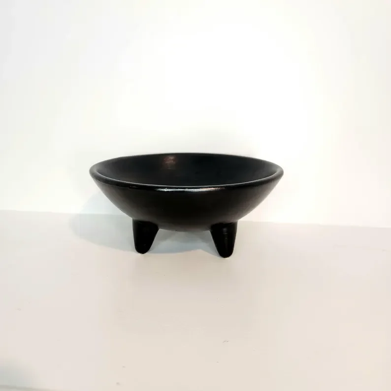A black ceramic bowl with three small legs, placed on a white surface against a white background.