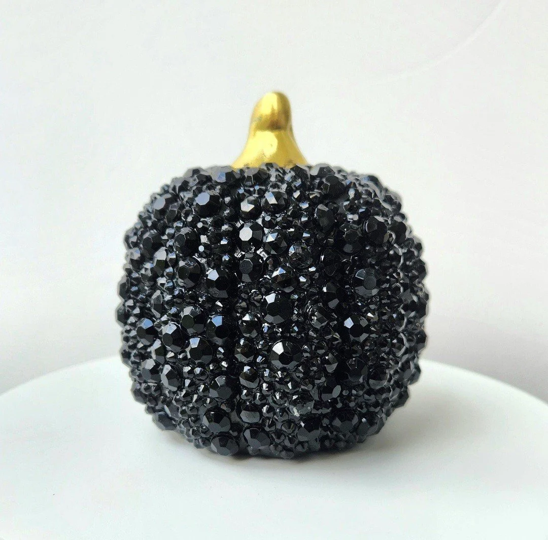 A decorative black berry ornament covered in small shiny black beads with a gold stem at the top, placed on a white surface against a plain white background.