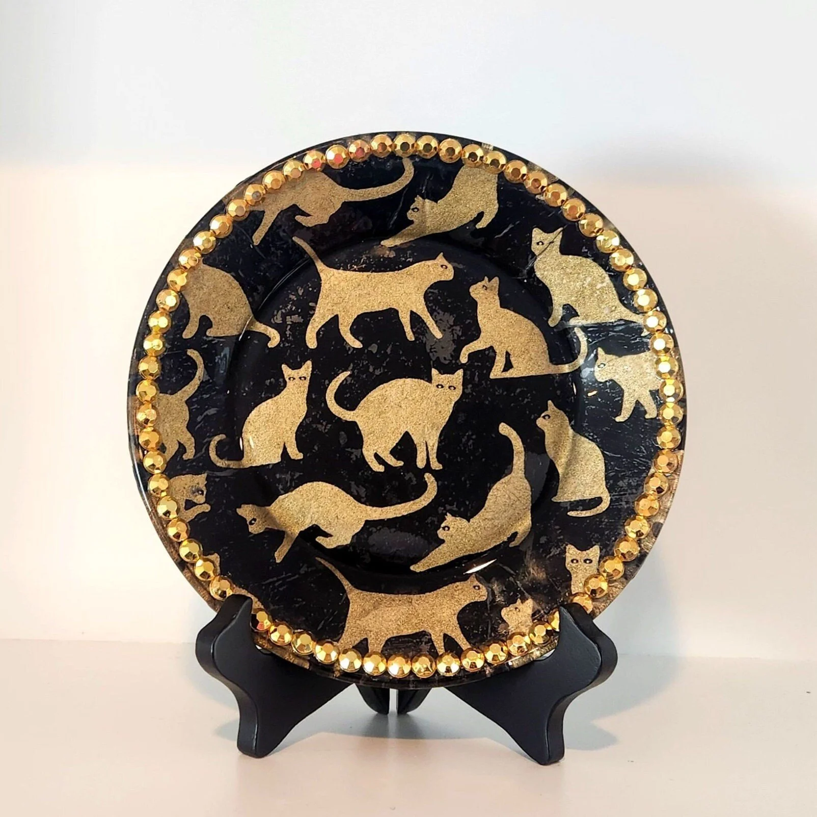 Decorative black plate with gold cat silhouettes and a border of gold beads, displayed on a black stand.