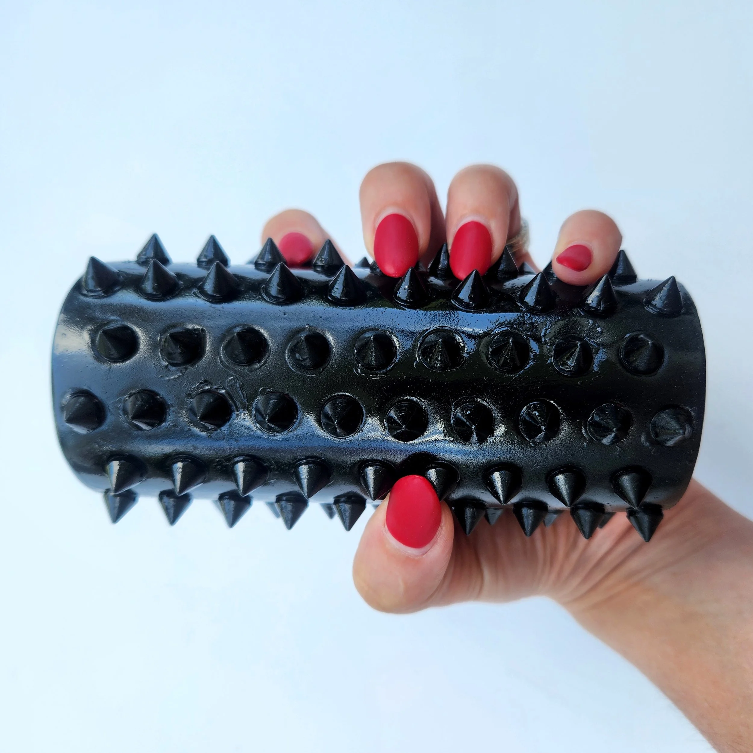 Hand with red-painted nails holding a black spiked roller massage tool against a white background.