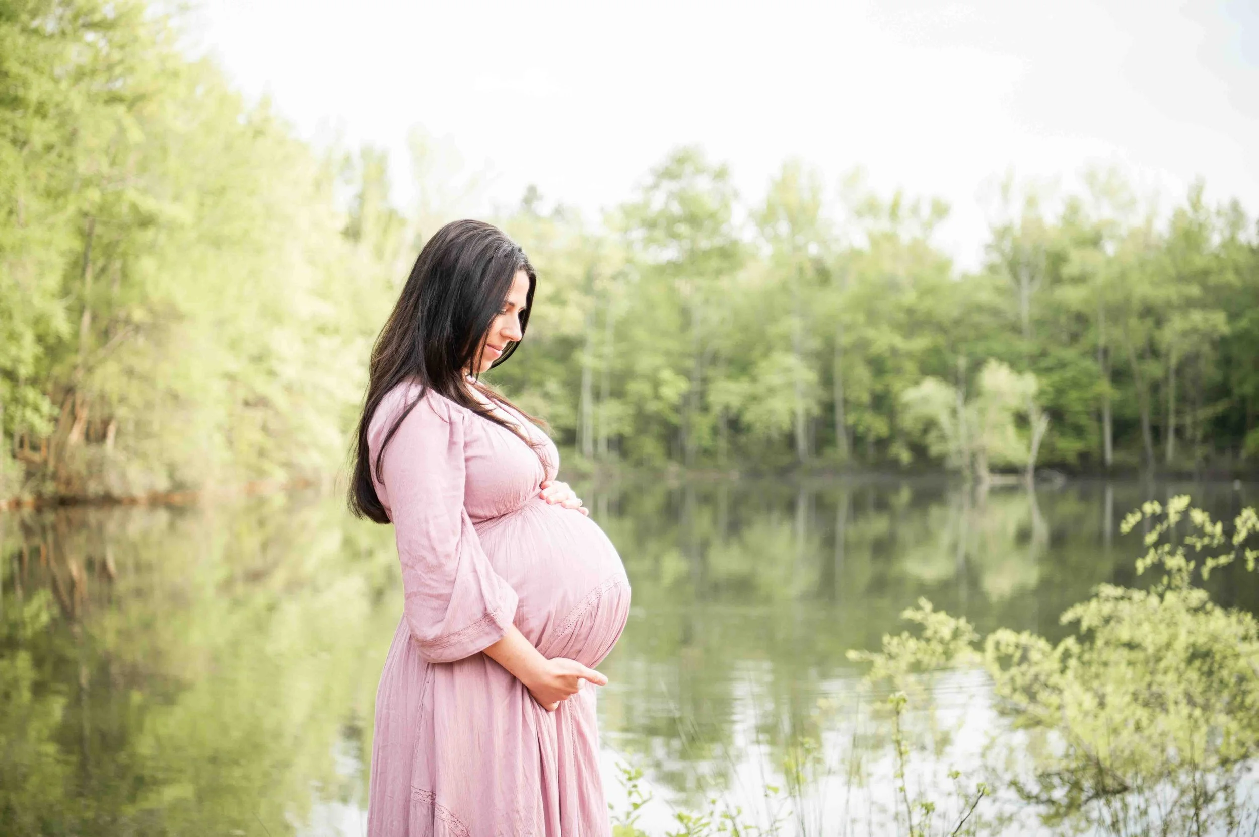 Pregnant woman in a pink dress stands beside a lake surrounded by green trees, holding her belly and looking down.