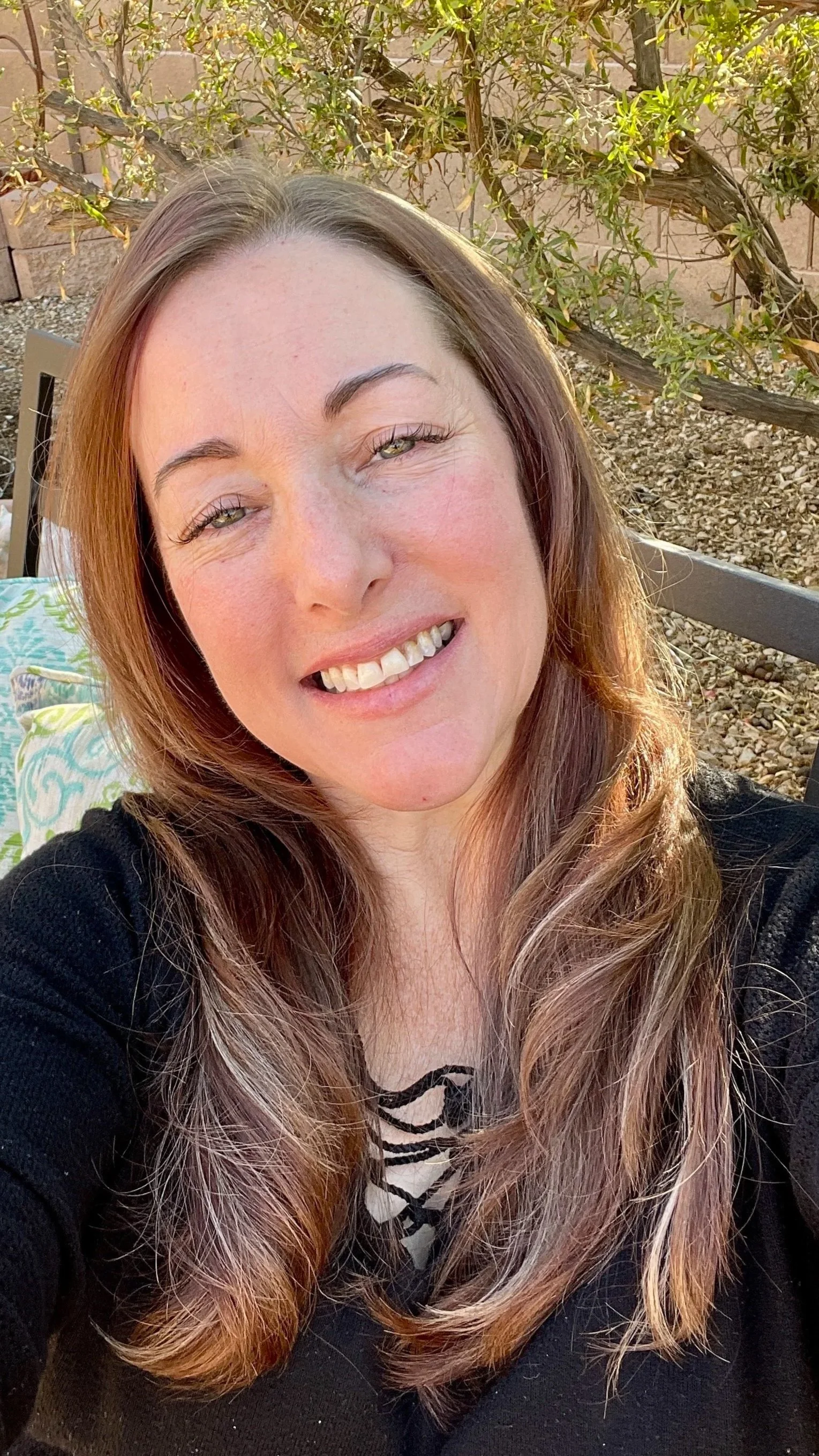 A woman with long, wavy brown hair and light skin takes a selfie outdoors. She is smiling with her teeth visible and has light-colored eyes. Behind her, there are bushes and a brick wall, with sunlight illuminating her face.