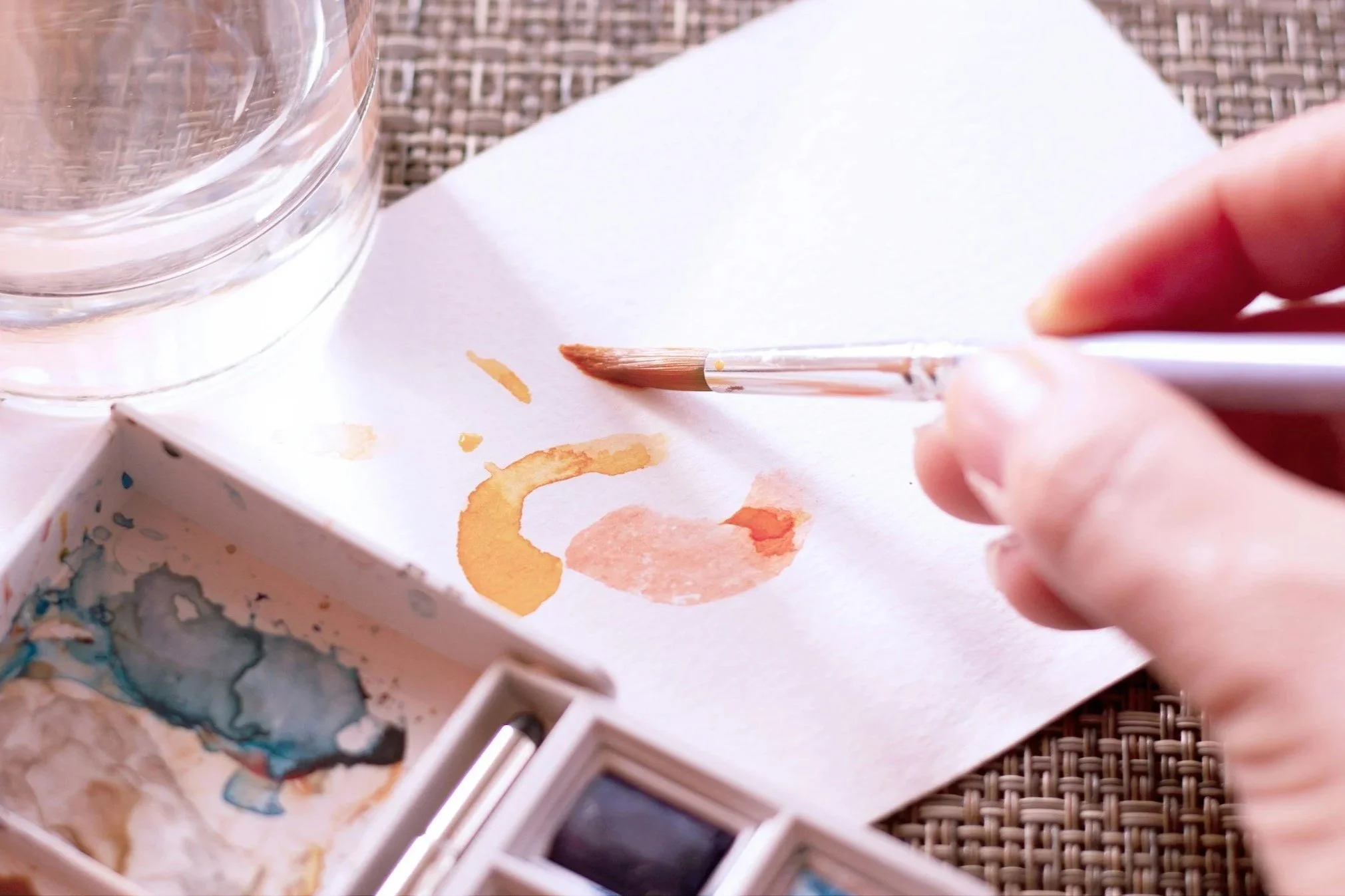Person painting with watercolors on paper, with watercolor palette and glass of water nearby.