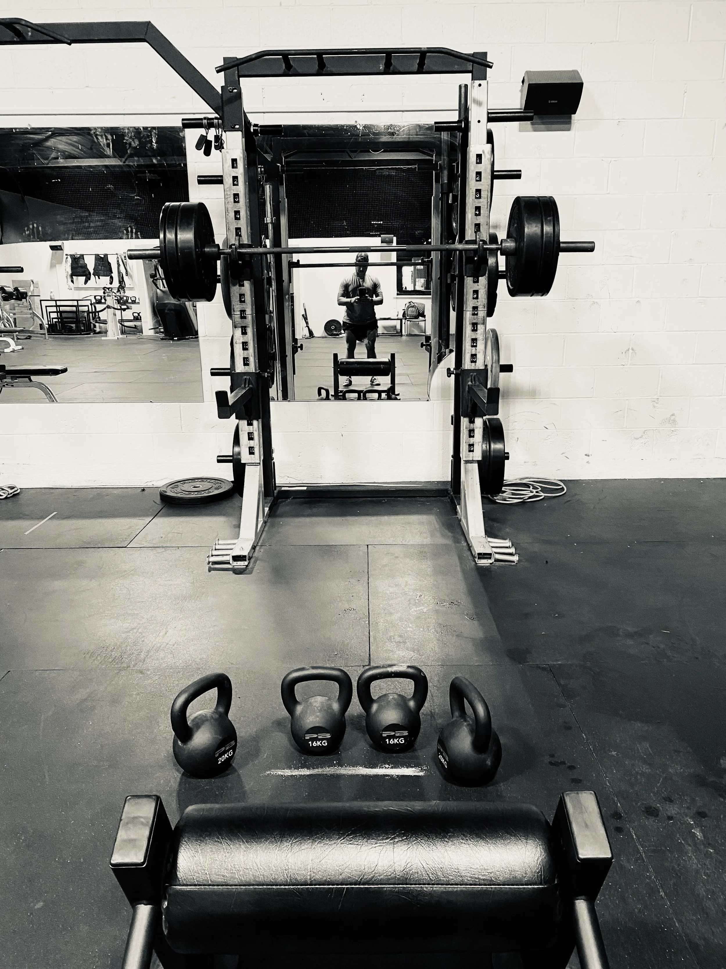 Weightlifting gym with a mirror reflecting a person taking a photo, kettlebells on the floor labeled 16 KG and 20 KG, and a power rack with weights.