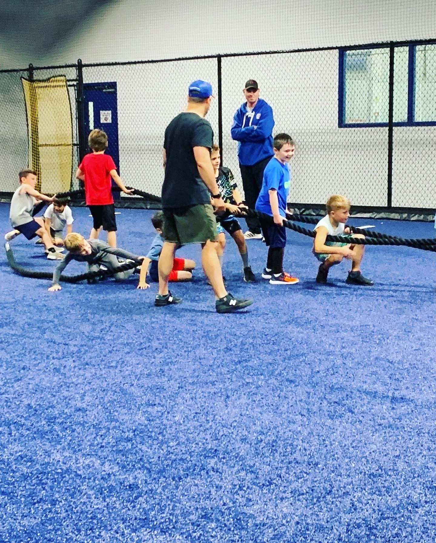 Children and an adult participating in a tug of war game indoors on a blue carpet, with a black fence and window in the background.