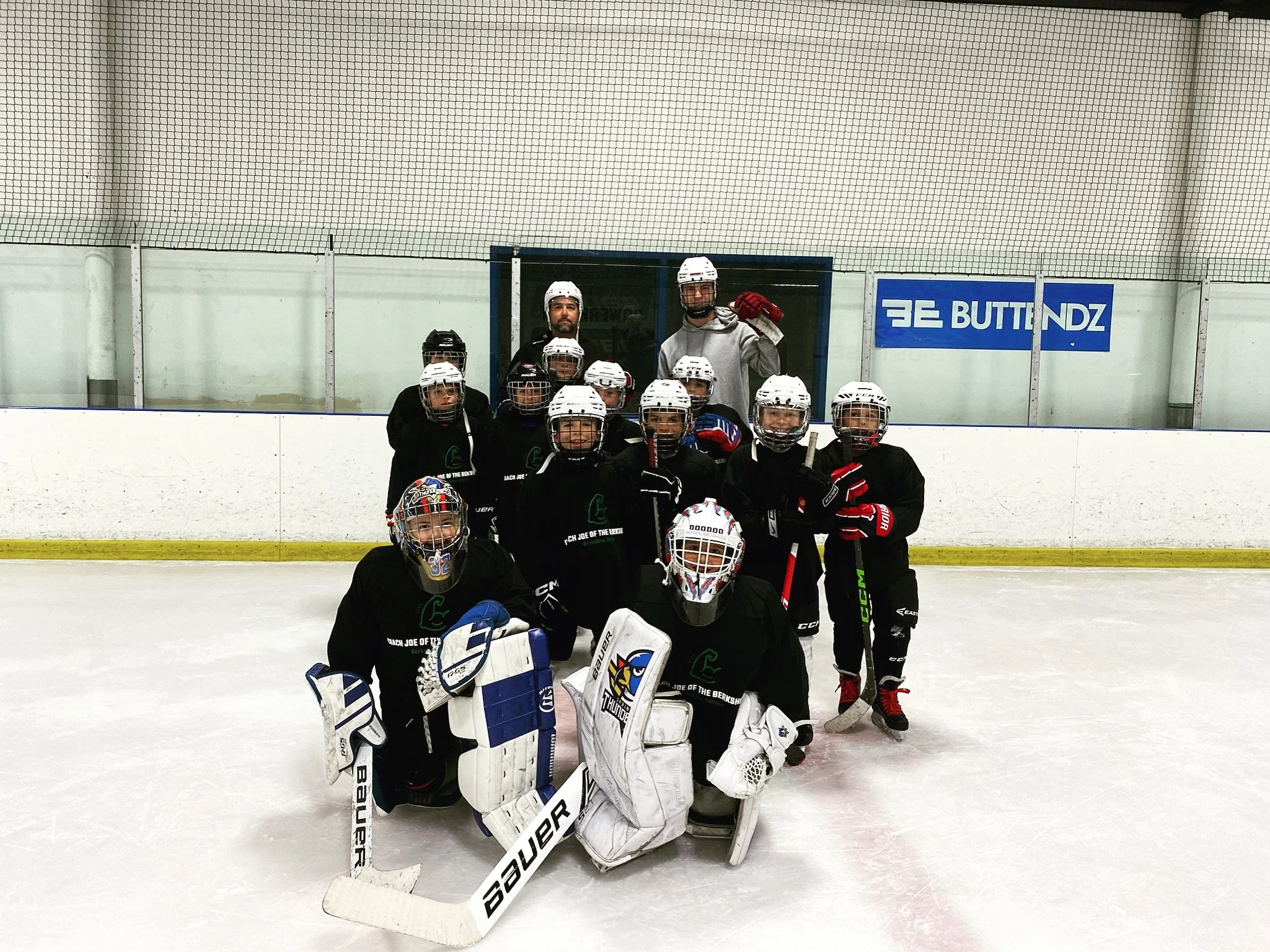 A group of young ice hockey players and their coaches posing on the ice rink, all wearing hockey gear including helmets and skates, with a coach holding a hockey stick and raising a fist.