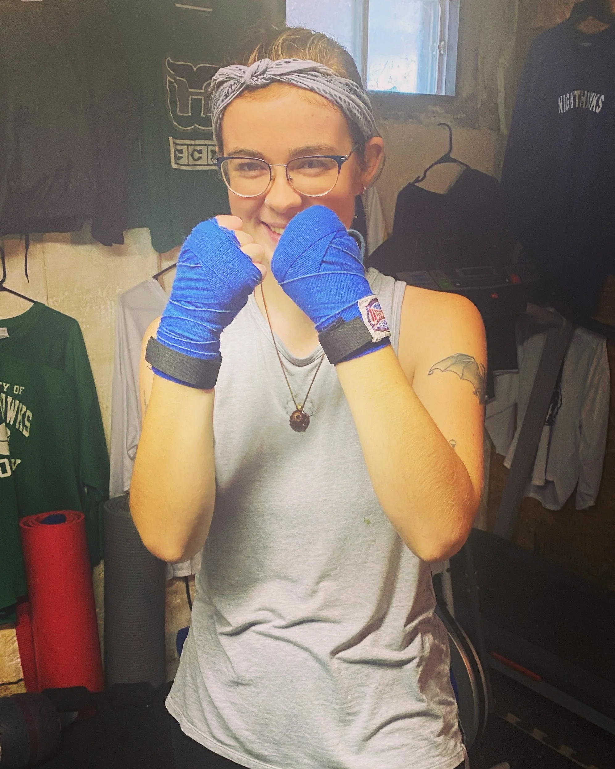 A woman with glasses and a bandana tied around her head, standing in a gym, smiling, wearing blue hand wraps, a sleeveless shirt, with tattoos on her arm, and boxing gloves hidden behind her hands.