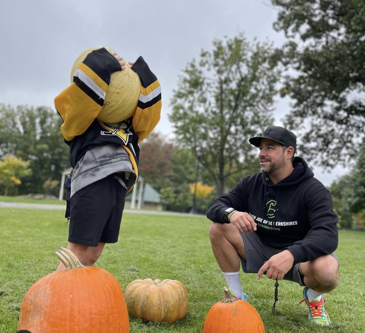 A man kneeling on the grass, smiling at a person holding a pumpkin over their head in a park with trees, during daytime. There are three pumpkins on the ground, and the person holding the pumpkin is wearing a sports jersey and shorts.