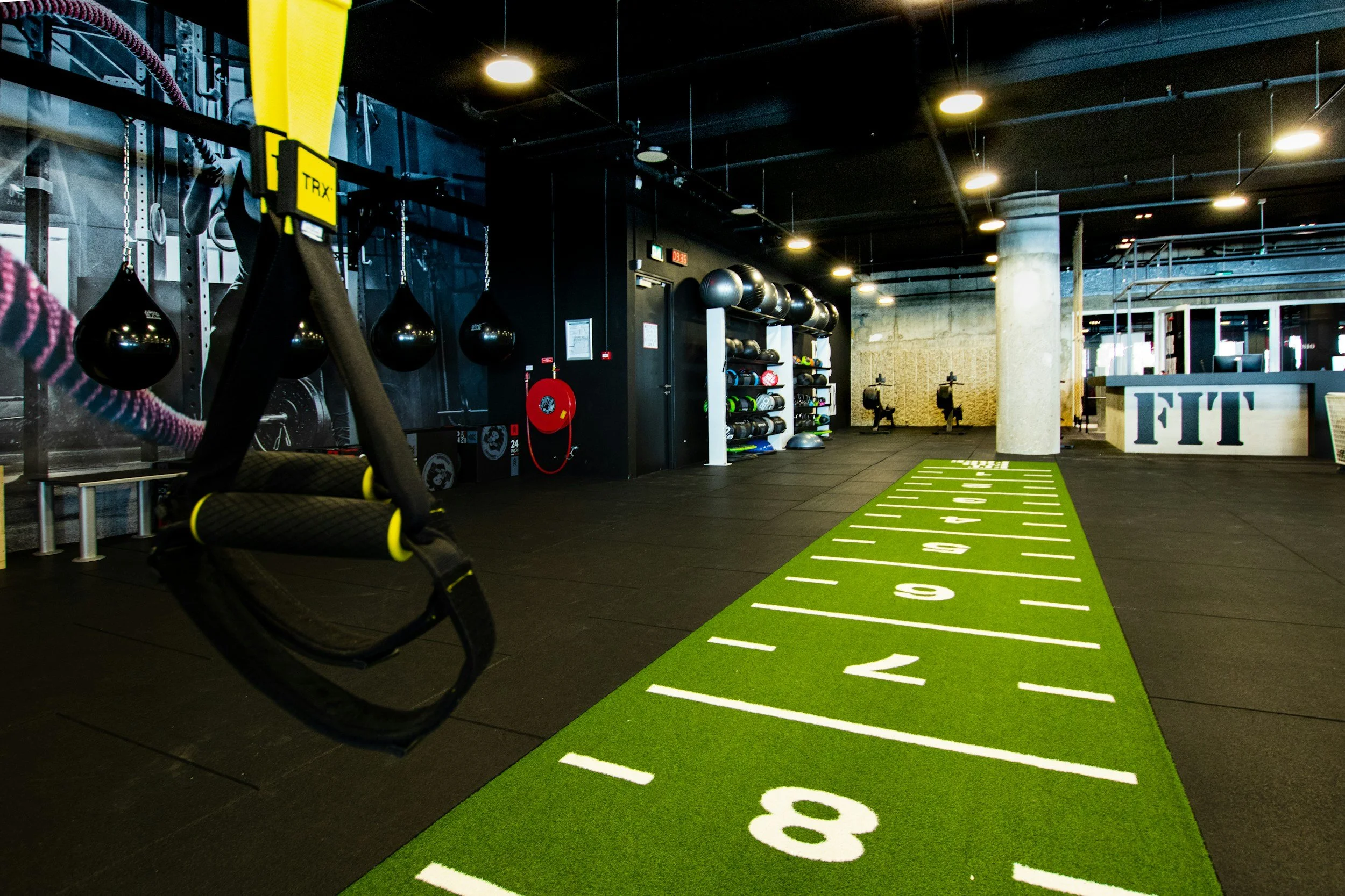 Indoor gym with black exercise equipment, hanging punching bags, wall-mounted weights, and a green running strip with numbered lanes from 1 to 8.