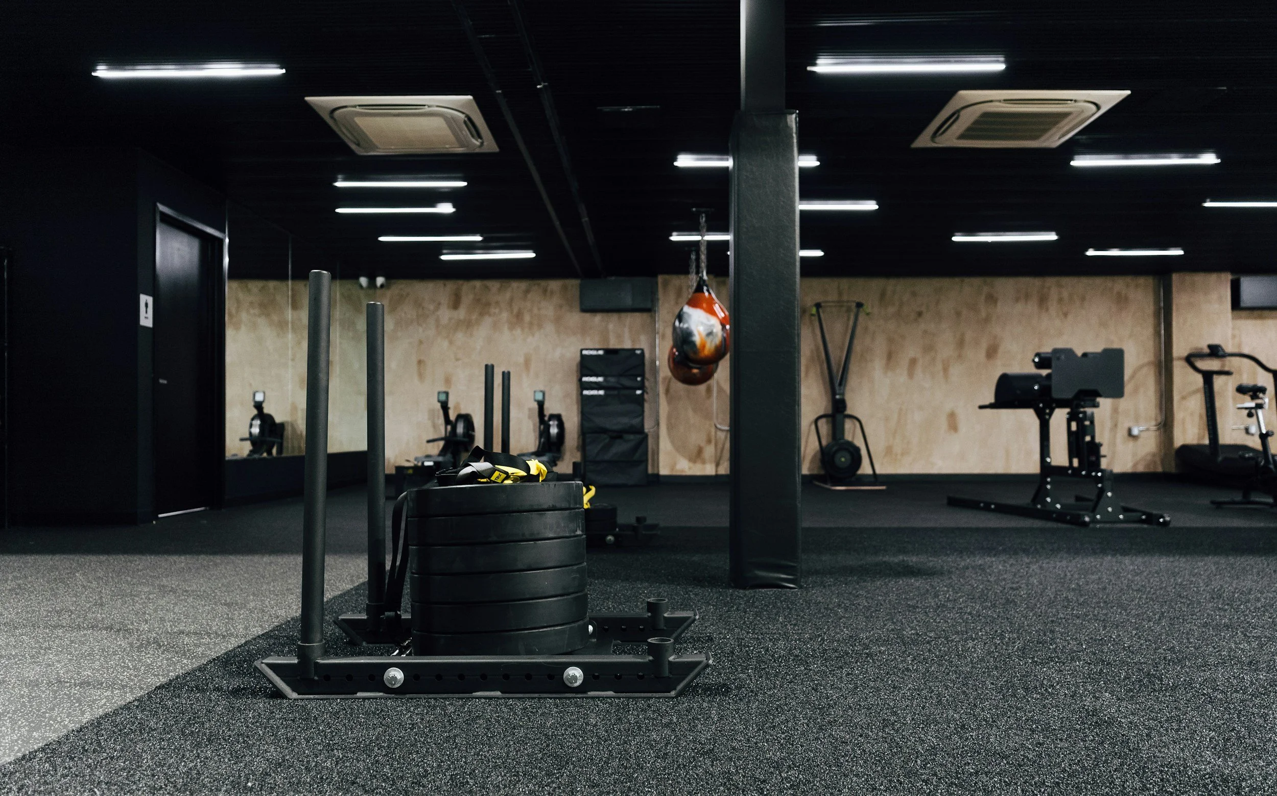 Empty gym with black rubber flooring, weight equipment, punching bag, and a black wall with wood paneling in the background.