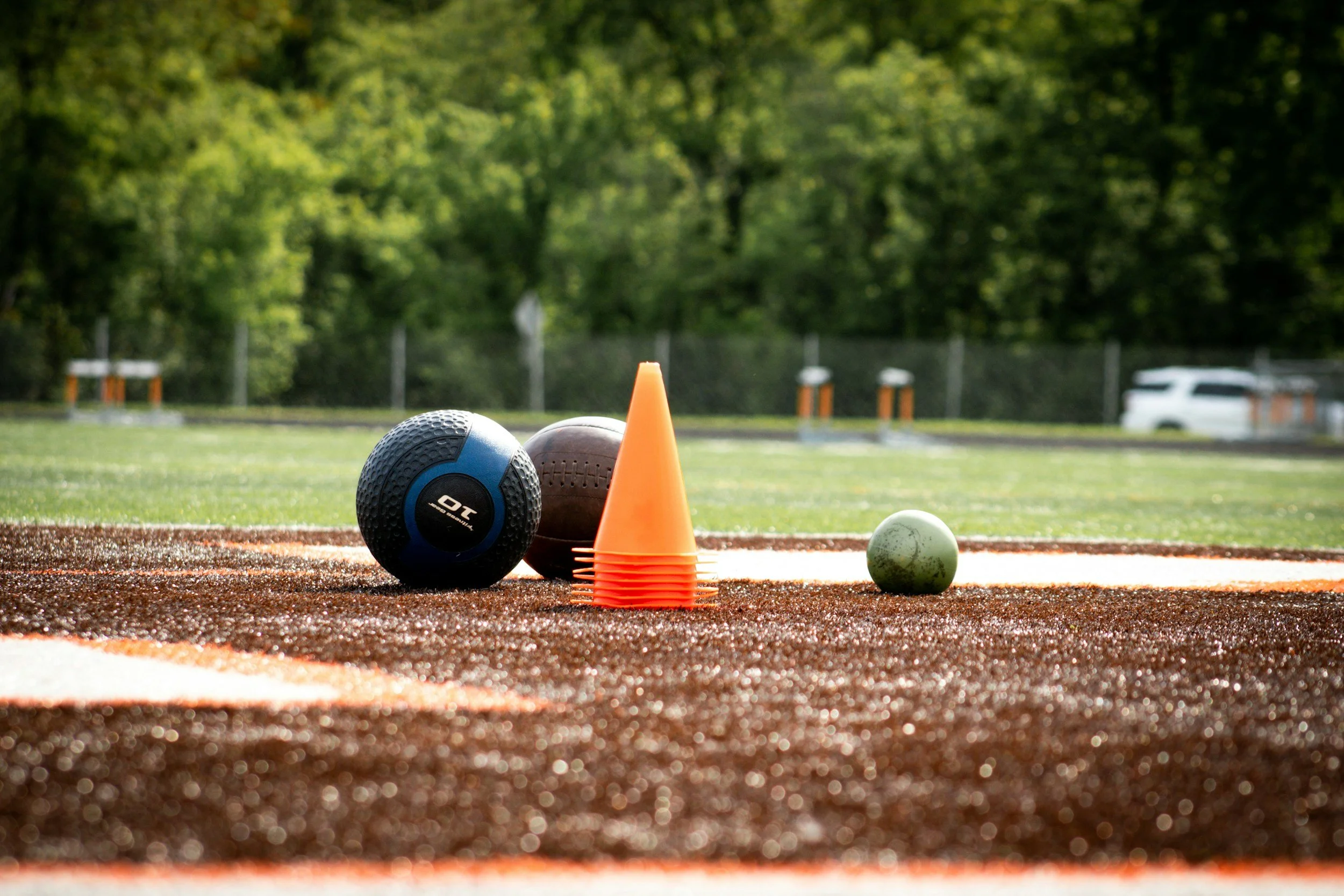 Sports equipment on a running track, including a blue medicine ball, a brown medicine ball, an orange cone, and a small white ball, with green trees and parked cars in the background.