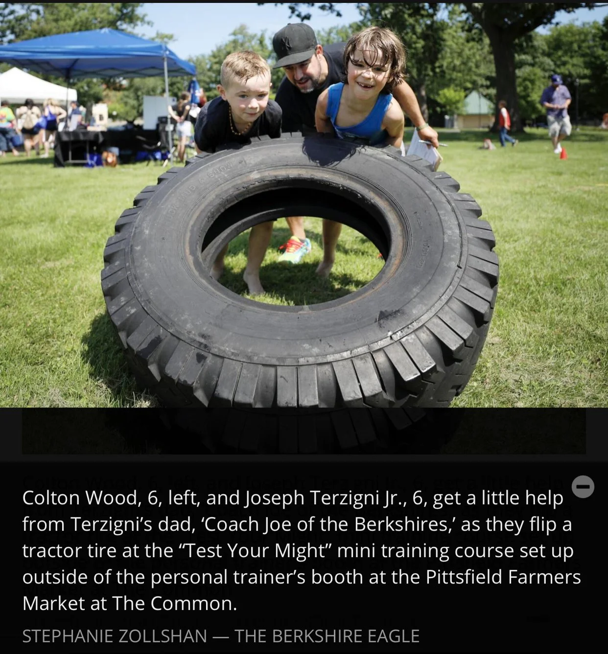 Two young boys, Colton Wood and Joseph Terzigni Jr., are leaning over a large tractor tire, with their hands on it, during an outdoor event. Their dad helps them with a mini training course at Pittsfield Farmers Market.