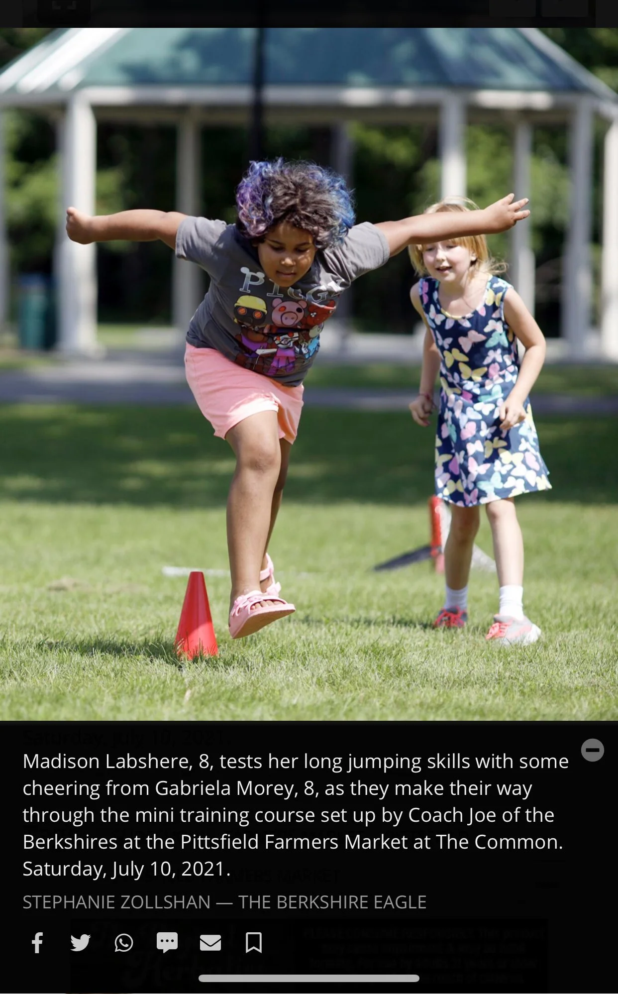 Two young girls are participating in a long jumping activity outdoors. One girl, with curly hair decorated with purple, is mid-air jumping over an orange cone, while the other girl, with blonde hair, is watching and smiling. There is a gazebo structu