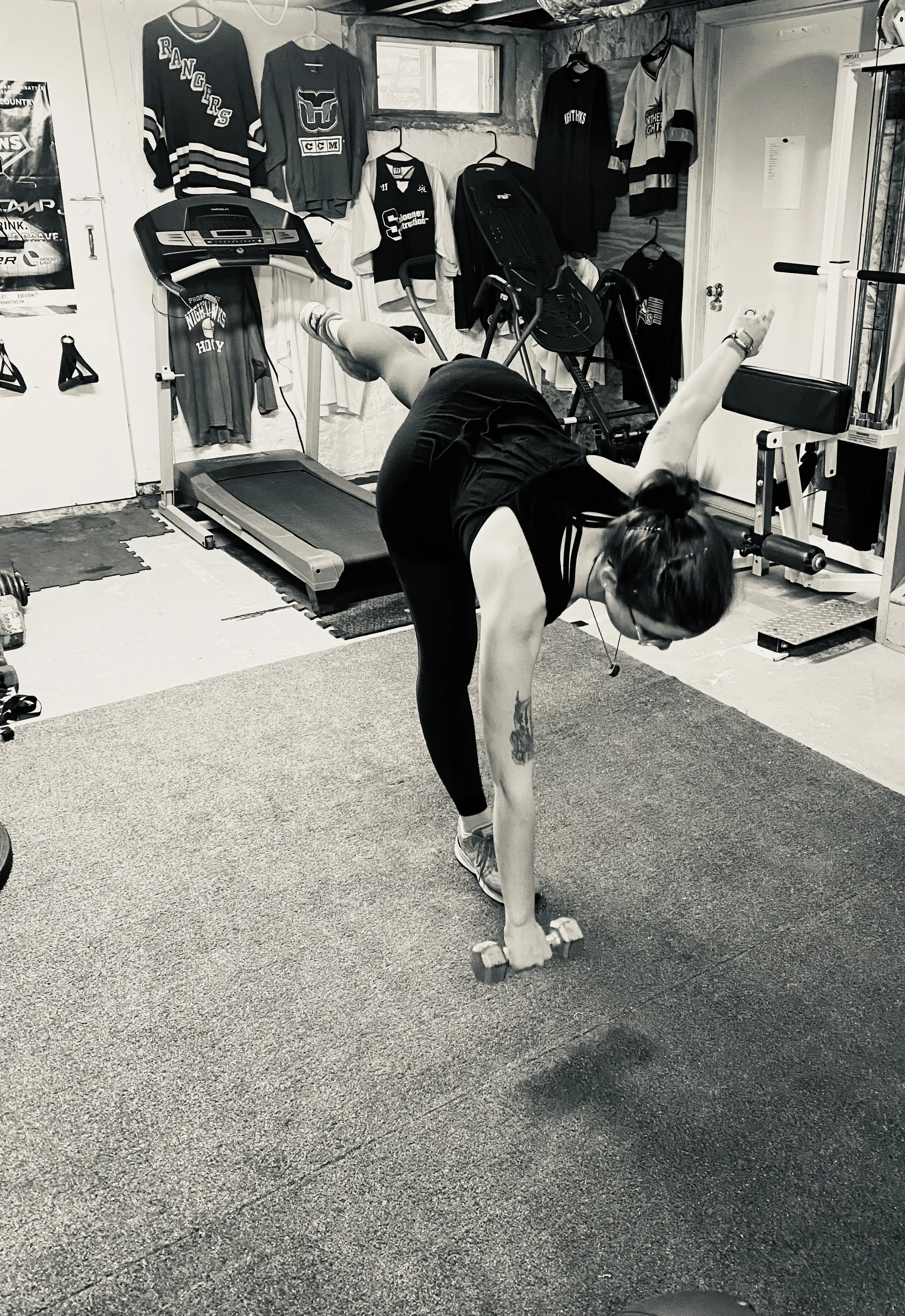 A woman in athletic clothing performing a forward bend stretching exercise in a home gym, with workout equipment and hockey jerseys hanging on the wall.