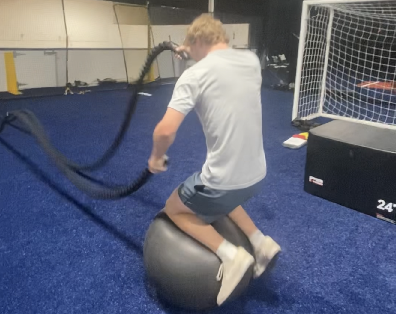 Young man performing battle ropes exercise over a Bosu ball in a gym with blue flooring and fitness equipment.
