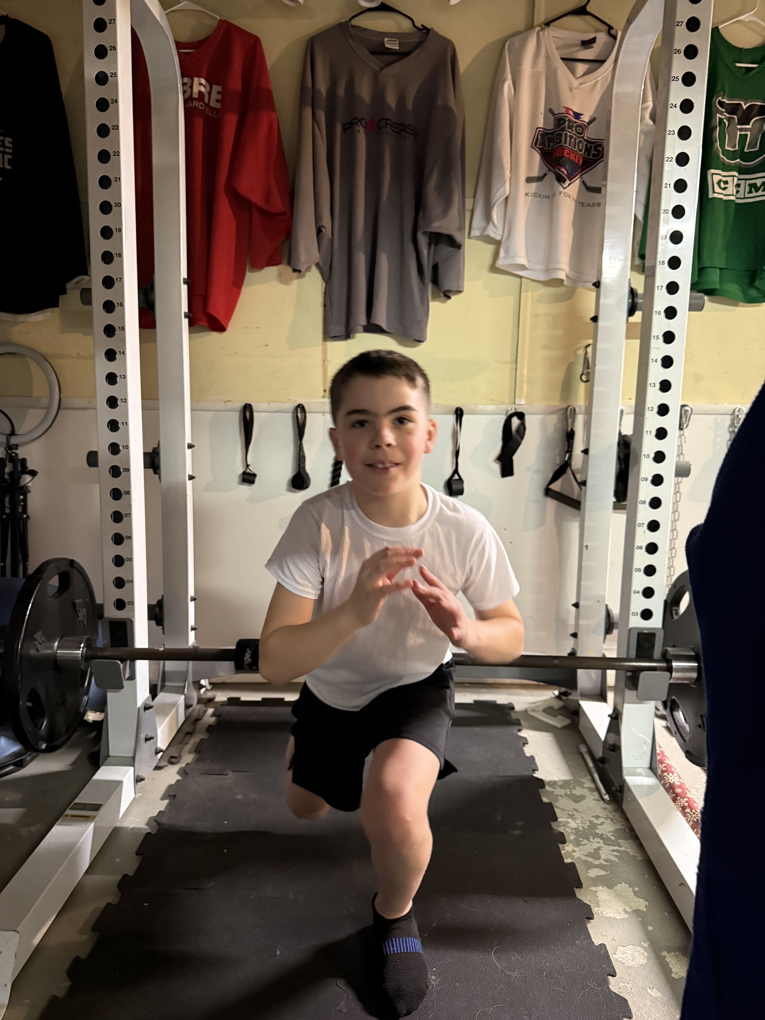A young man in a white t-shirt and black shorts is doing a lunge exercise in a home gym, with weightlifting equipment and hanging shirts in the background.