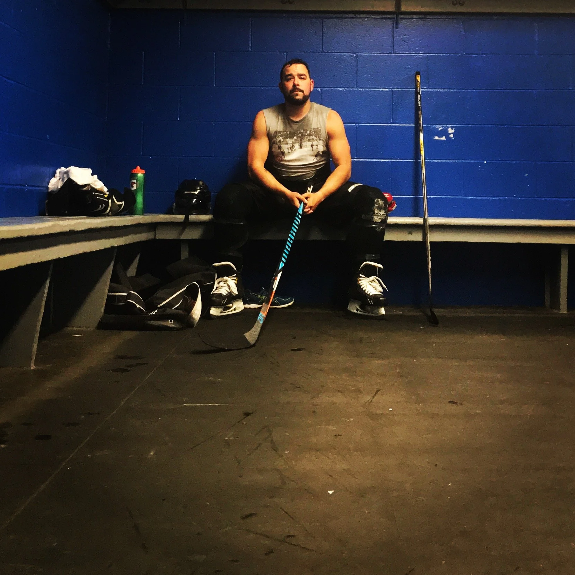A man sitting on a bench in front of a blue wall, holding a hockey stick, with sports gear and water bottle around him. A hockey stick is propped up against the wall beside him.