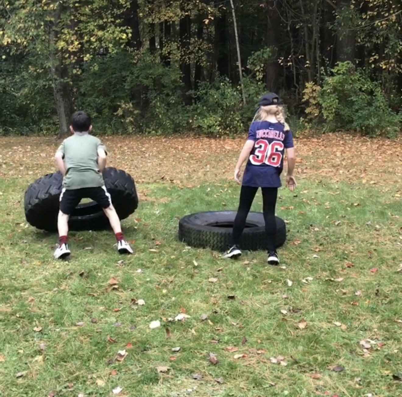 Two children outdoors in a grassy area, one boy shirtless and wearing shorts, lifting a large tire, and a girl with long hair, wearing a sports jersey and a cap, standing near a tire on the ground. There are trees and autumn leaves in the background.