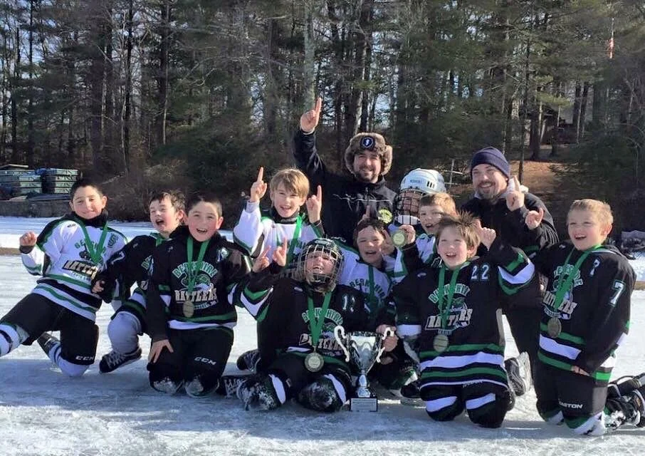 Youth hockey team with medals and coaches, celebrating on the ice after winning a game, outdoors with trees in the background