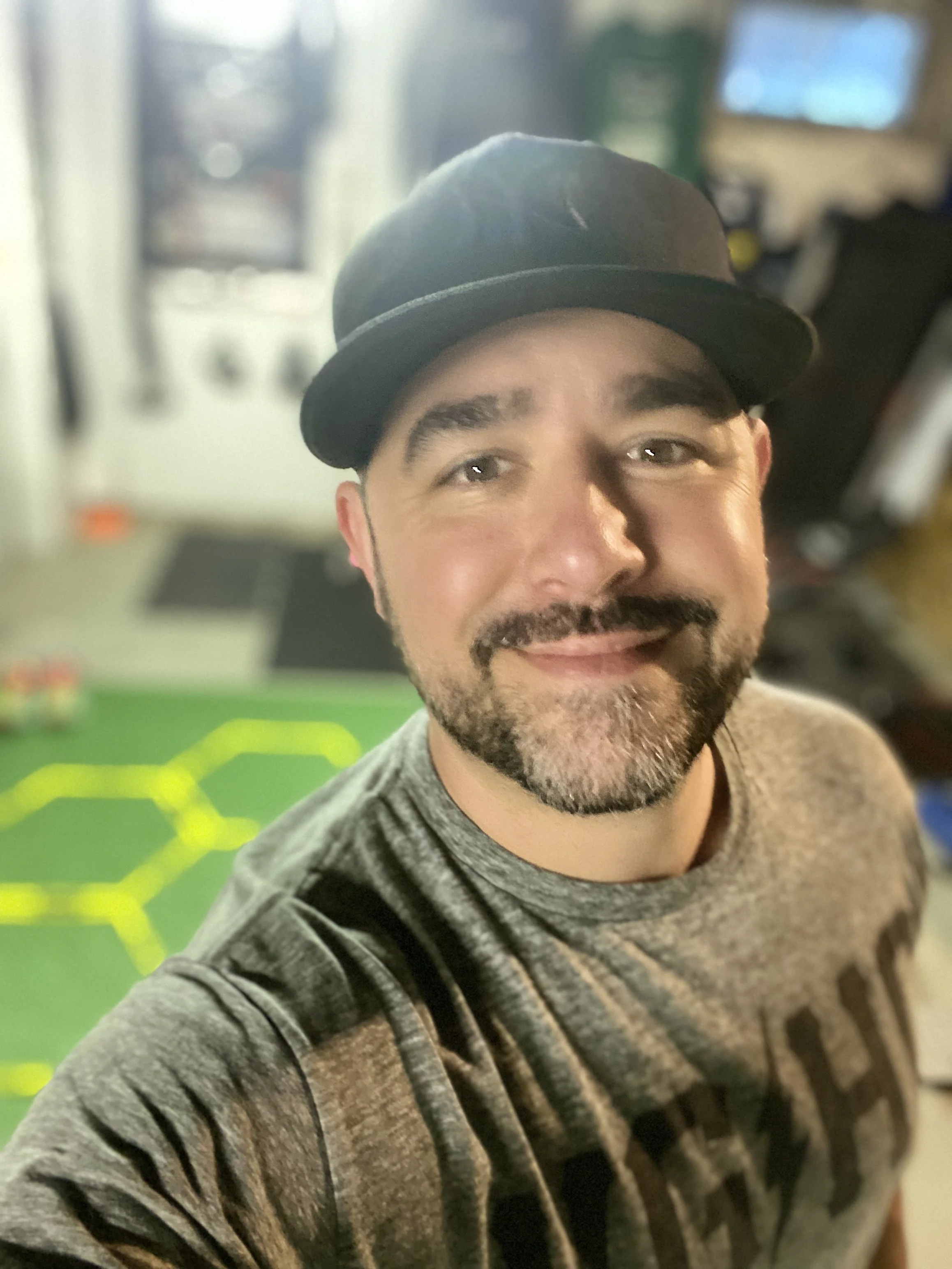 A man smiling wearing a gray shirt and a black hat, with a blurred background that includes a green surface with yellow markings and a vending machine.