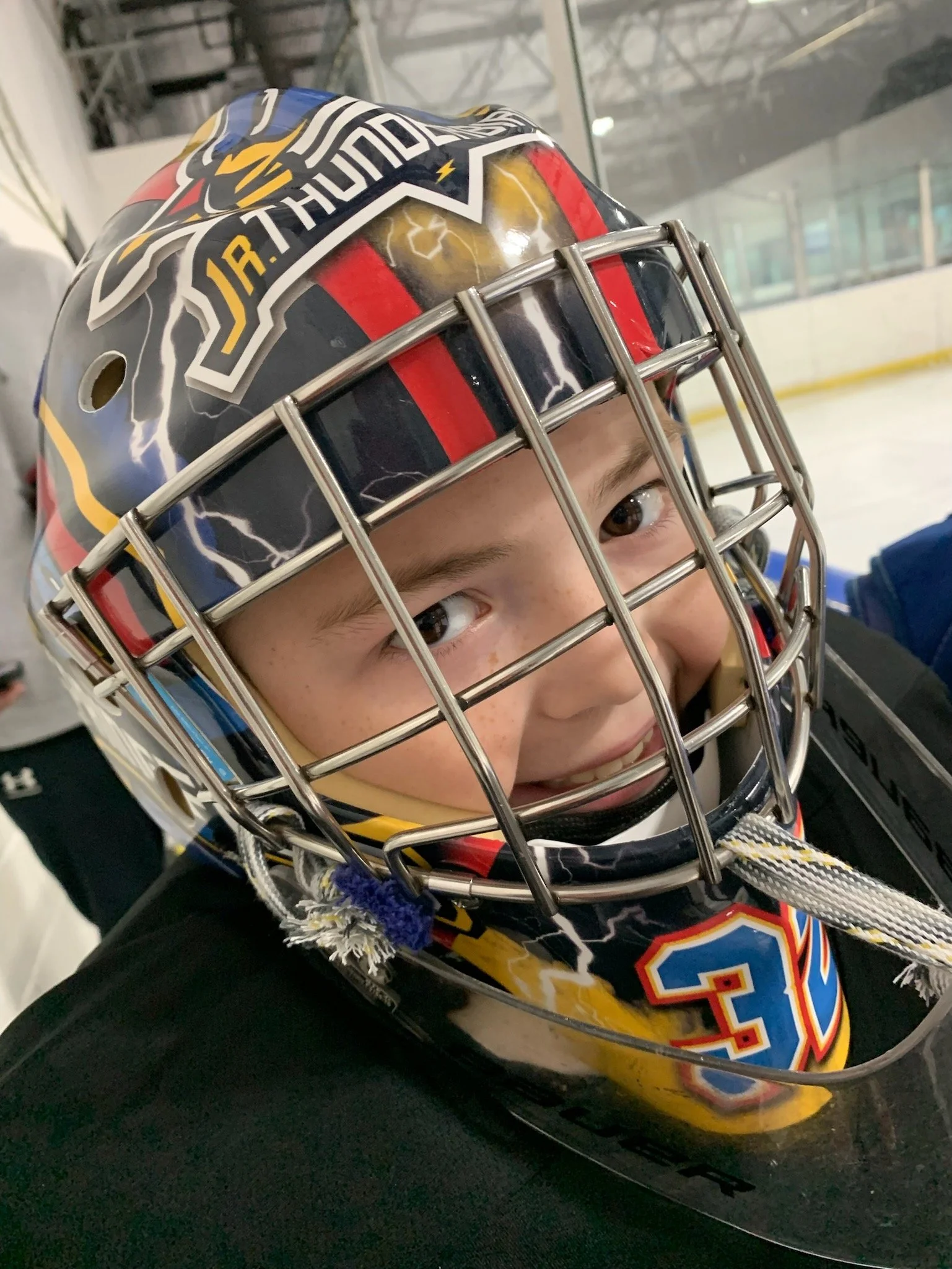 Close-up of a young hockey player wearing a helmet with a cage, featuring lightning and lightning bolt designs, and a jersey with the number 34. The player is on an ice rink.