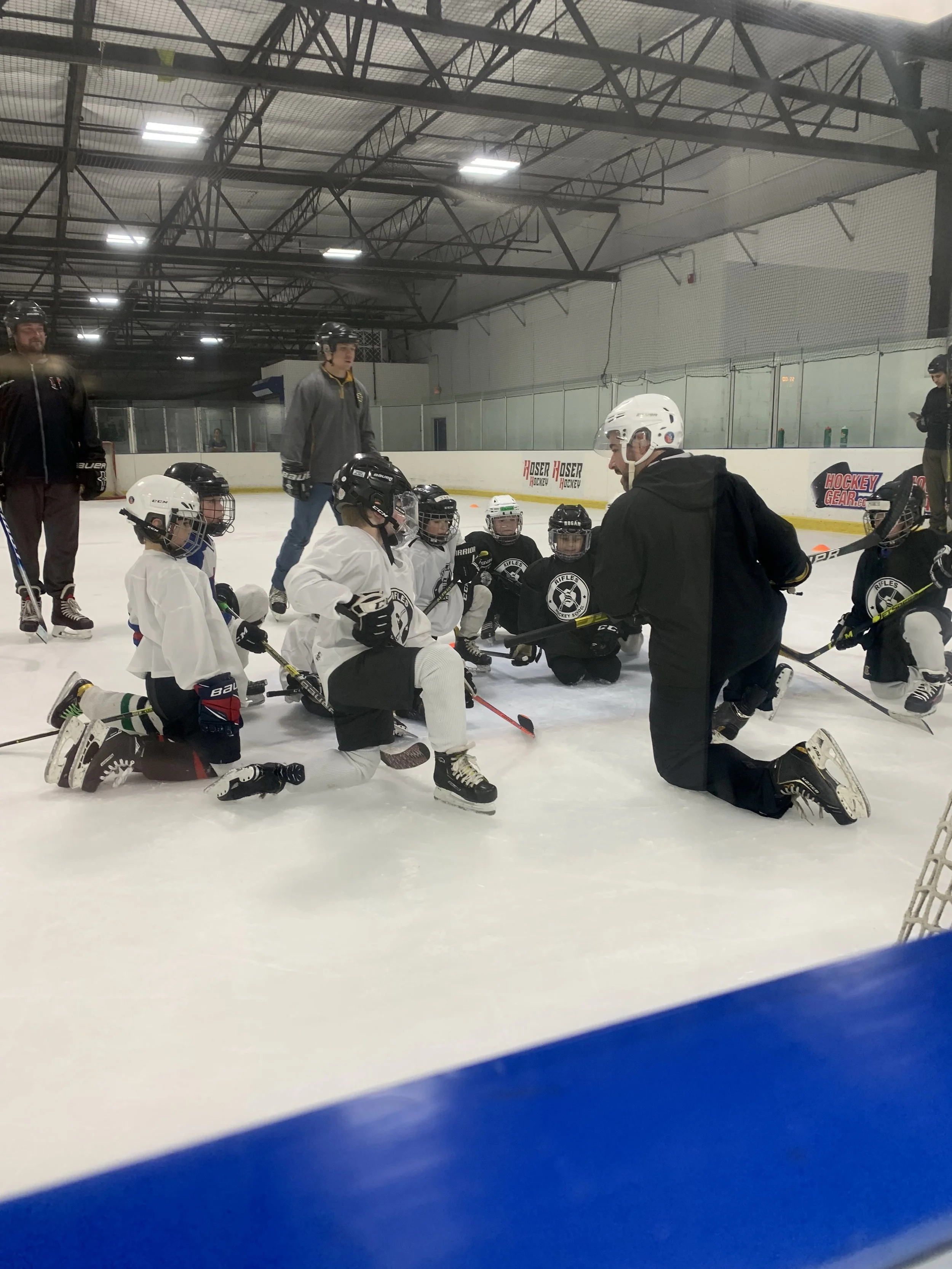 Group of young hockey players in helmets and jerseys sitting on the ice rink floor during a coaching session, with their coach kneeling and giving instructions.